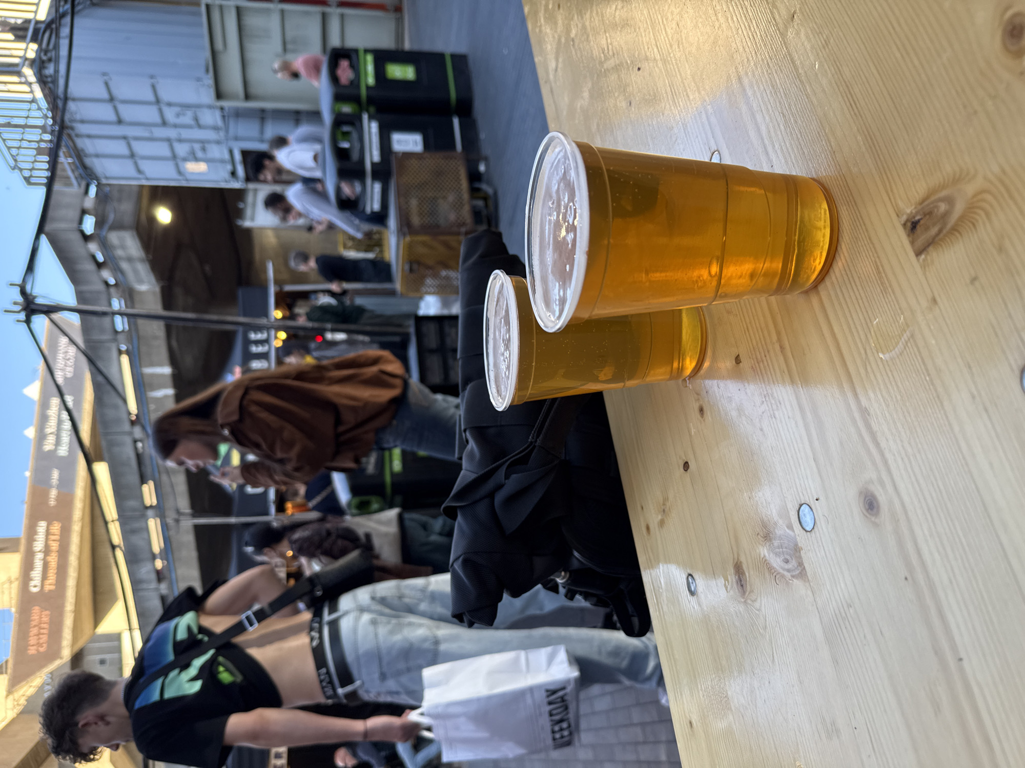 The scene depicts an outdoor gathering with a focus on two plastic cups filled with a yellow beverage, likely beer, resting on a light-colored wooden table. Behind the cups, several individuals are milling about. One individual, a young male, is prominently featured in the foreground, wearing a black t-shirt with green and blue accents, light-wash jeans, and a black cross-body bag. He is carrying a white shopping bag with "WEEKDAY" printed on it. Further in the background, other people are engaged in conversations and activities. Behind these people is a structure with the writing “Chiharu Shiota Threads of life HAYWARD GALLERY” written on it. The general environment is urban, possibly a street food or market area. The location is suggested to be in London, United Kingdom.