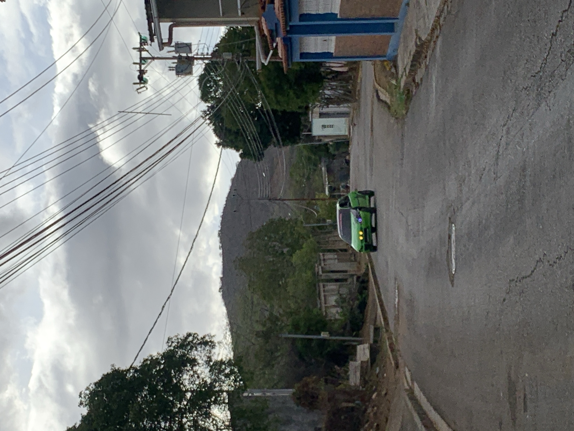 The image captures a daytime scene in La Tagua, Venezuela, under an overcast sky filled with gray clouds. The primary subject is a paved road that curves gently to the right, showing visible cracks and wet patches on its surface.
A bright green car, possibly a sedan or small SUV, is positioned on the right side of the road, facing away from the viewer, with its front lights discernible. To the left, numerous utility poles are visible, supporting a complex web of electrical or communication wires that crisscross against the sky.
The background features a prominent hill or mountain, sparsely covered with vegetation, suggesting a rugged or dry landscape. Various trees and dense foliage are present throughout the scene, particularly along the road and at the base of the hill. On the right side of the road, some structures are visible, including one with blue and reddish-brown elements, and a white box-like structure that appears to have some faint, illegible black text on its side. No people are visible, and no specific activity is occurring. The scene conveys a quiet, semi-rural or outer urban environment.