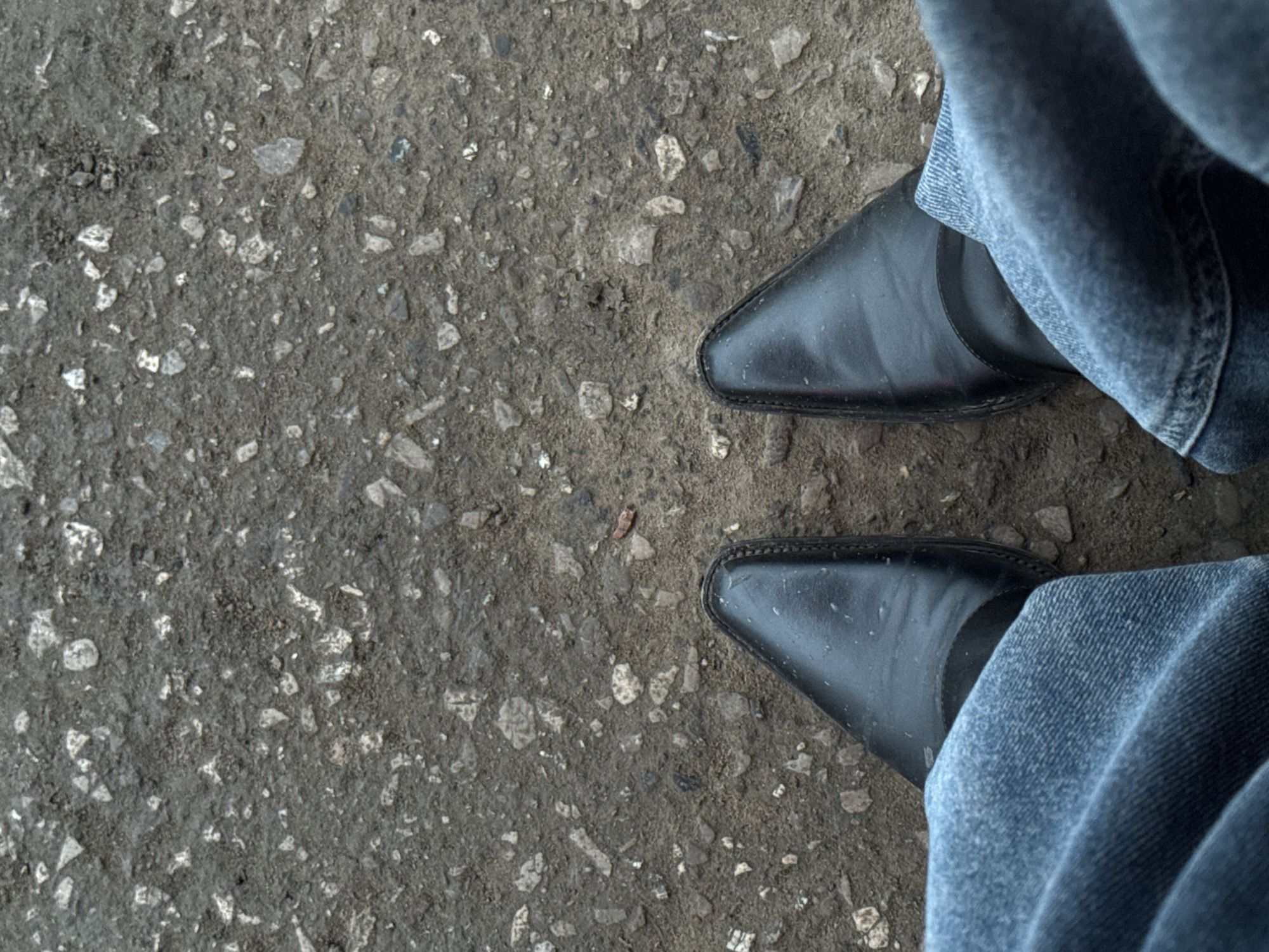 The image depicts a view from above, focusing on a section of rough, gray asphalt ground. Two black, pointed-toe shoes are positioned near the right side of the frame, partially covered by blue jeans. The asphalt surface is textured with small stones and uneven patches. The setting appears to be a typical urban environment.