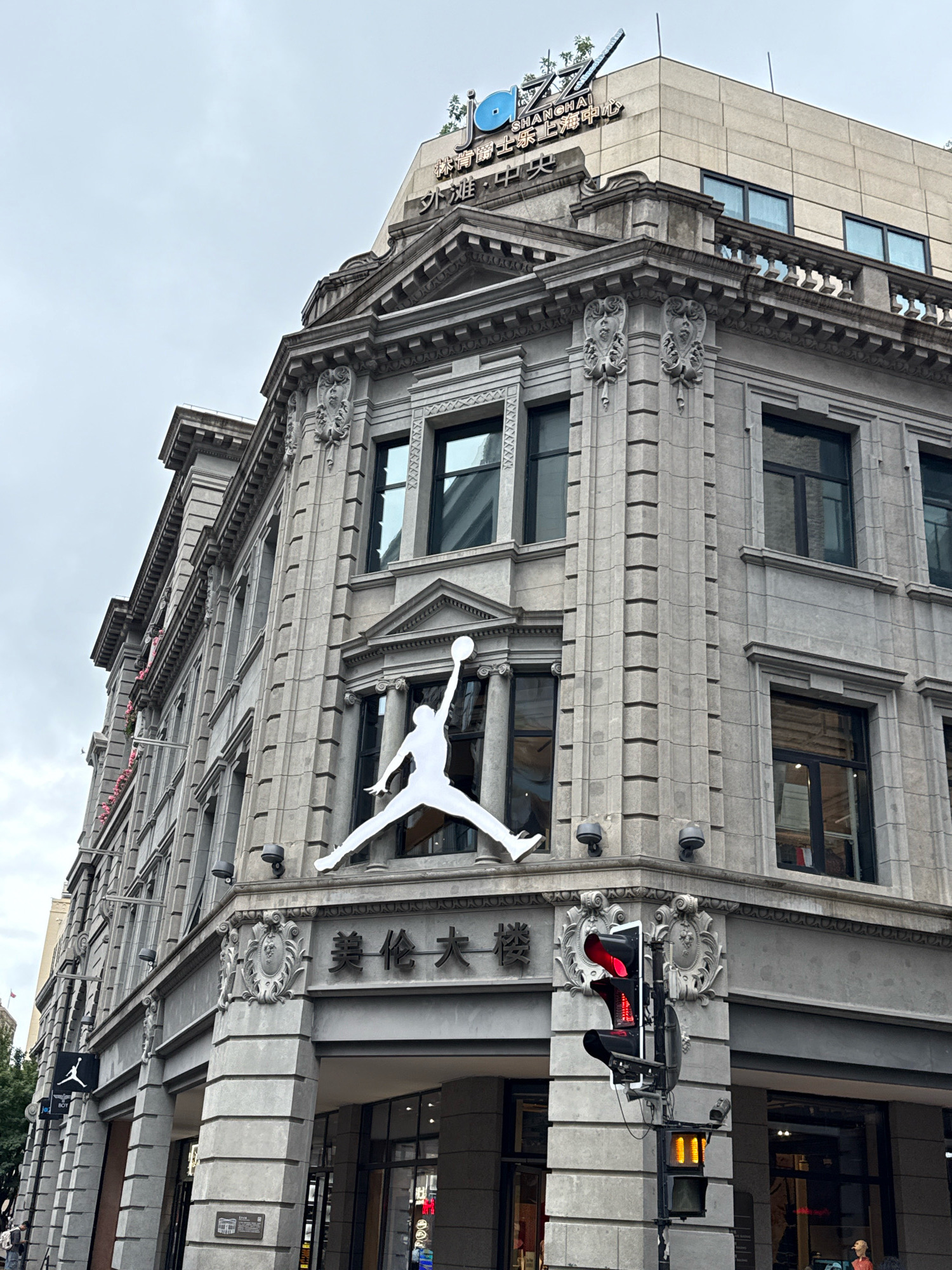 This image captures the exterior of a multi-story building in Shanghai, China. The building features classical architectural elements, including ornate stonework, columns, and decorative carvings. A prominent white silhouette of the iconic Jumpman logo, associated with the Nike brand, is displayed on the corner of the building. Above this, the Chinese characters "美伦大楼" are visible. Higher up on the building, a sign with the word "JAZZ" in a stylized font and Chinese characters indicating "SHANGHAI" and "CENTER" is illuminated. A traffic light showing a red signal is positioned in the foreground, attached to a pole. The sky is overcast.