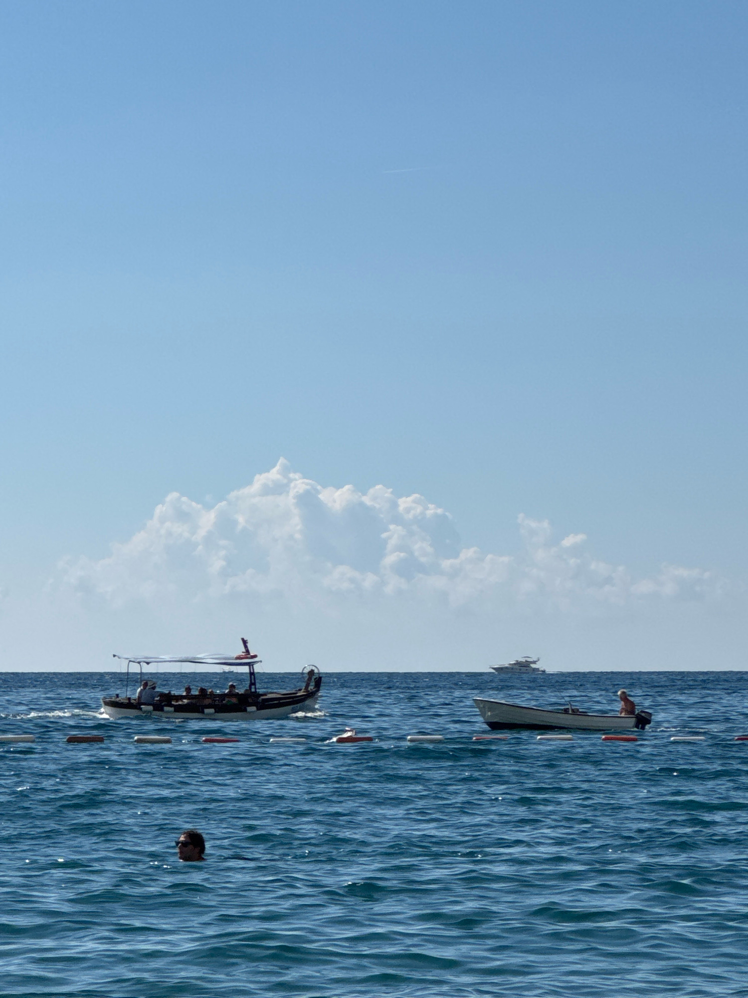 The image captures a scene on the waters off Boreti, Montenegro, under a clear blue sky with scattered white clouds. In the foreground, the deep blue sea is rippled by gentle waves. A person's head and shoulders are visible above the water, wearing sunglasses. A row of buoys marks a boundary in the water.
Further out, a long, narrow boat with a canopy is carrying several passengers. Behind this boat, a smaller white motorboat is visible with a single person on board. In the distance, a large white yacht is sailing on the horizon. The overall impression is one of a peaceful day at sea, with various watercraft and individuals enjoying the coastal environment.