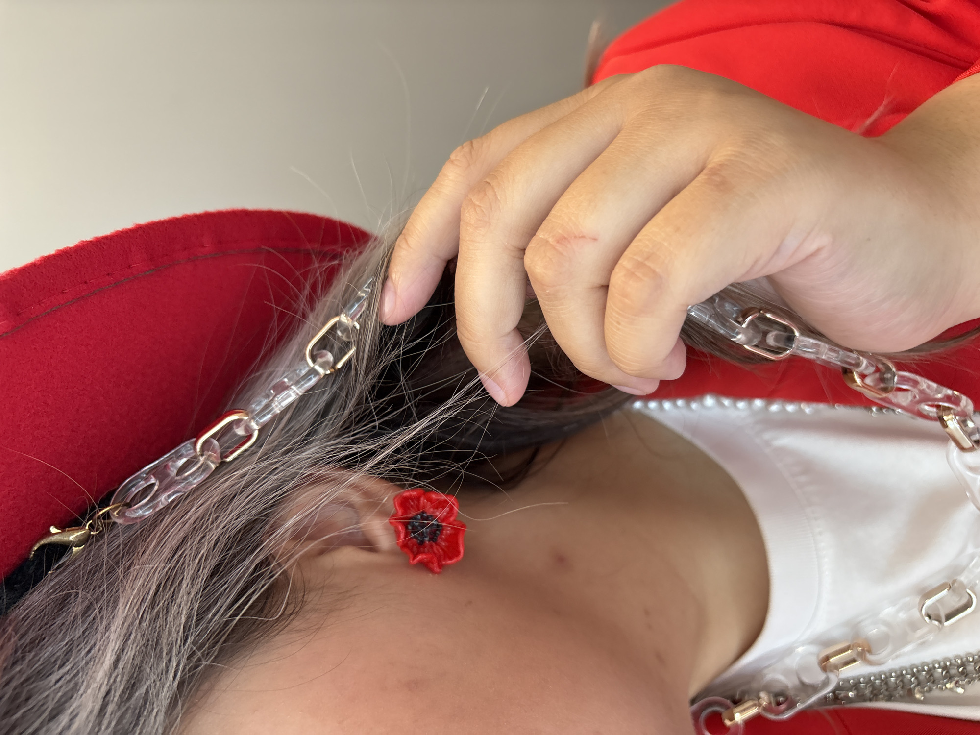A person's head and hand are depicted close-up. The person has graying hair, and is wearing a red hat and what appears to be a white shirt under a red jacket. A clear plastic chain with gold-colored links is draped across their hair. An earring shaped like a red poppy is visible. The person's hand is near their hair, and a small scratch is visible on the index finger. The background is a plain, light-colored wall. There is no information in the image to suggest it was taken in Birobidzhan, Russia.