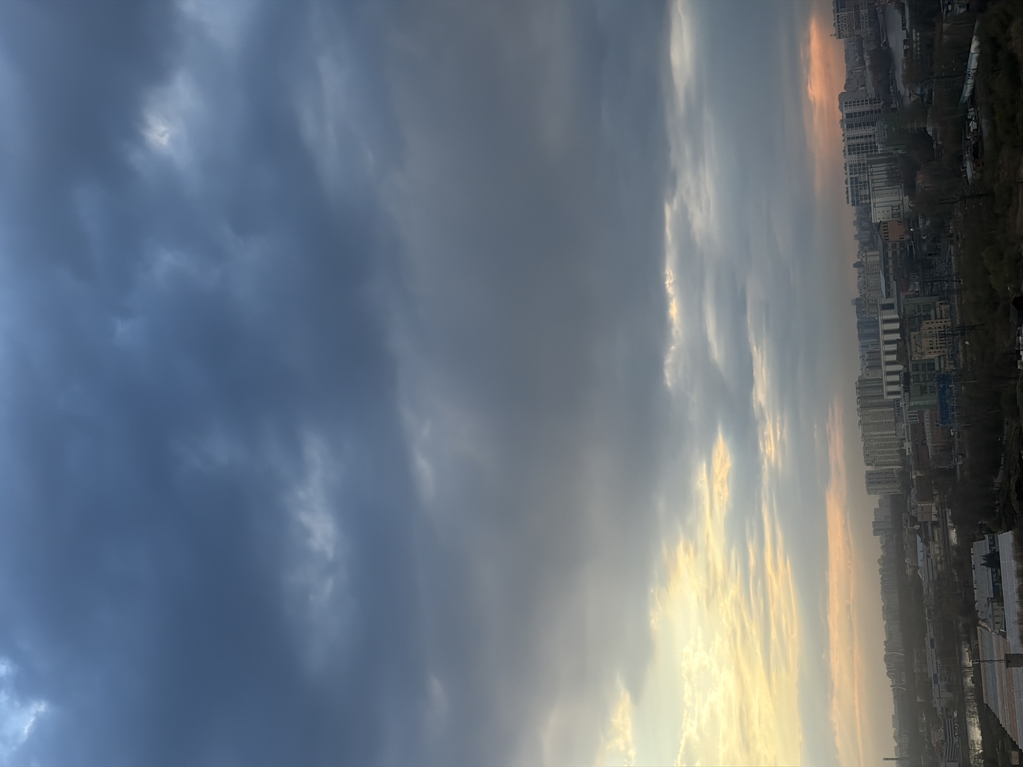 The image displays a wide panoramic view of a cityscape under a dramatic sky, captured during either sunrise or sunset in Saint Petersburg, Russia. The sky occupies the majority of the frame, exhibiting a striking contrast. On the left side, dense, dark grey and blue-grey storm clouds dominate, creating a heavy and somewhat brooding atmosphere. In stark contrast, the right side of the sky is illuminated by warm, golden, and orange light, indicating the presence of the sun breaking through or setting behind lighter clouds. Streaks of sunlight are visible through the cloud layers on this side.
Below the horizon, which is positioned in the lower portion of the image, lies a sprawling urban environment. Numerous multi-story buildings, appearing as residential blocks and commercial structures, stretch across the landscape. The cityscape is dense, with a mix of architectural styles, and some areas hint at industrial zones or possibly the banks of a river or canal. Dark, bare trees are sparsely visible along the edges of the city, suggesting early spring or late autumn.
The time of day is twilight, characterized by the low-angle, warm sunlight that creates the vibrant hues in the clouds. The weather is cloudy with partial clearing, leading to a dramatic interplay of light and shadow across the sky. No specific human activity or identifiable landmarks are visible. There is no visible text in the media file.