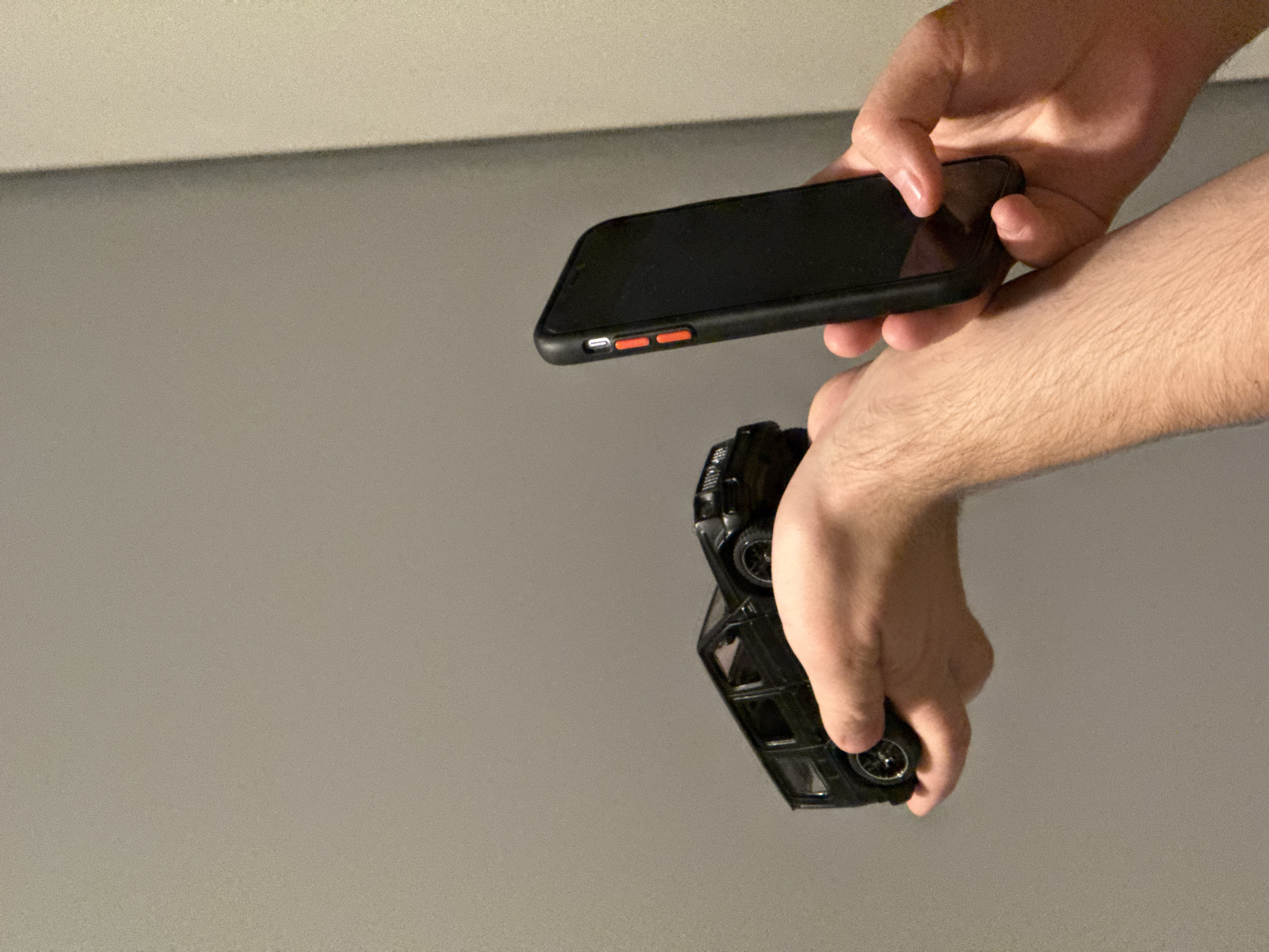 A person is holding a black toy car in their left hand and a black smartphone in their right hand, with the phone being held vertically. The background is a plain, textured wall with a shadow visible on the right side. The image does not provide any information about the city of Łódź, Poland.