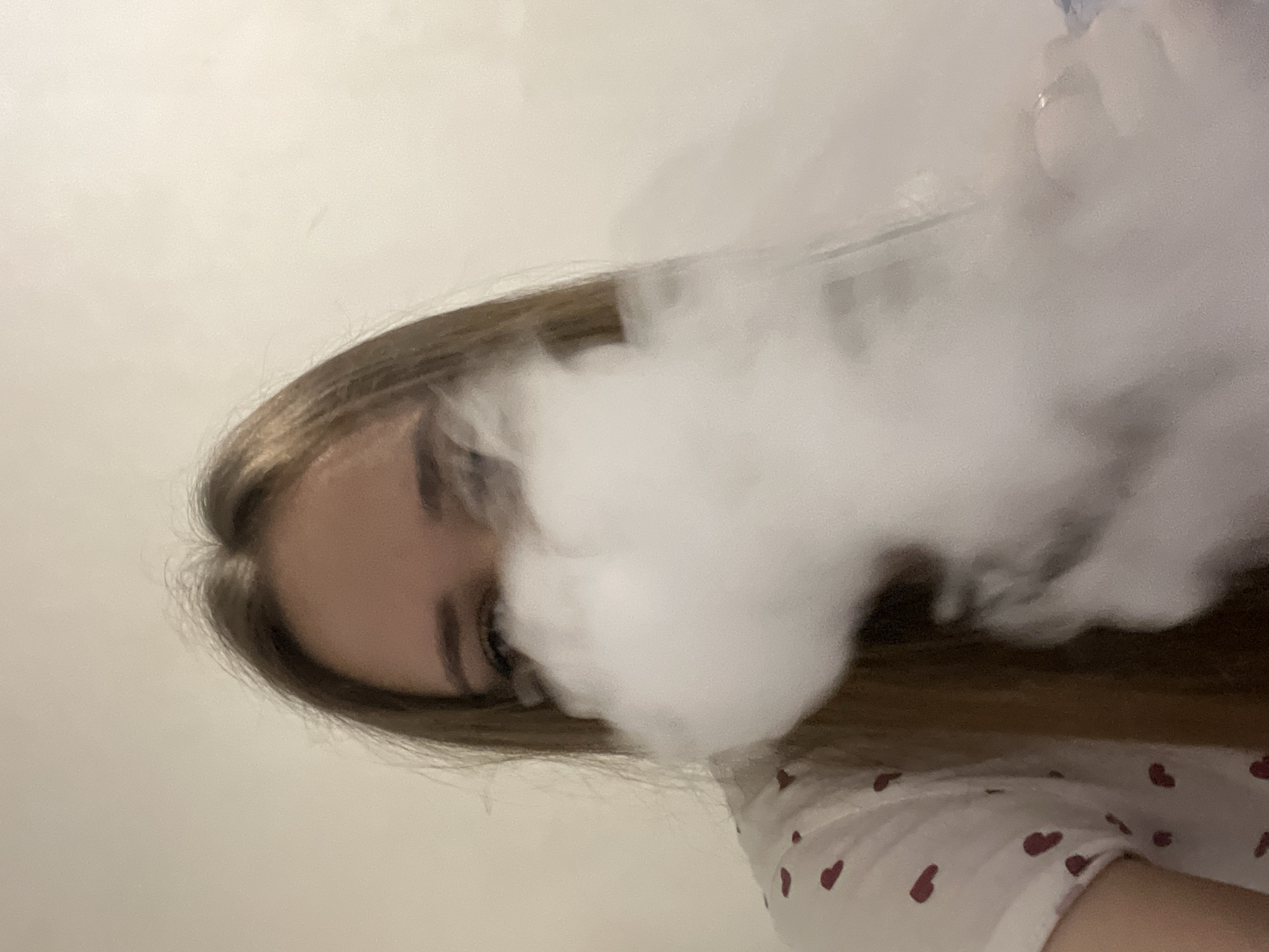A young woman with long, light brown hair is partially obscured by a large cloud of white vapor, possibly from a vape or some other smoking device. Only one of her eyes is clearly visible. She is wearing a white shirt with small red heart patterns. The background is a plain, light-colored wall. The image appears to be taken indoors, likely in a residential setting. Location is not discernible from the image.