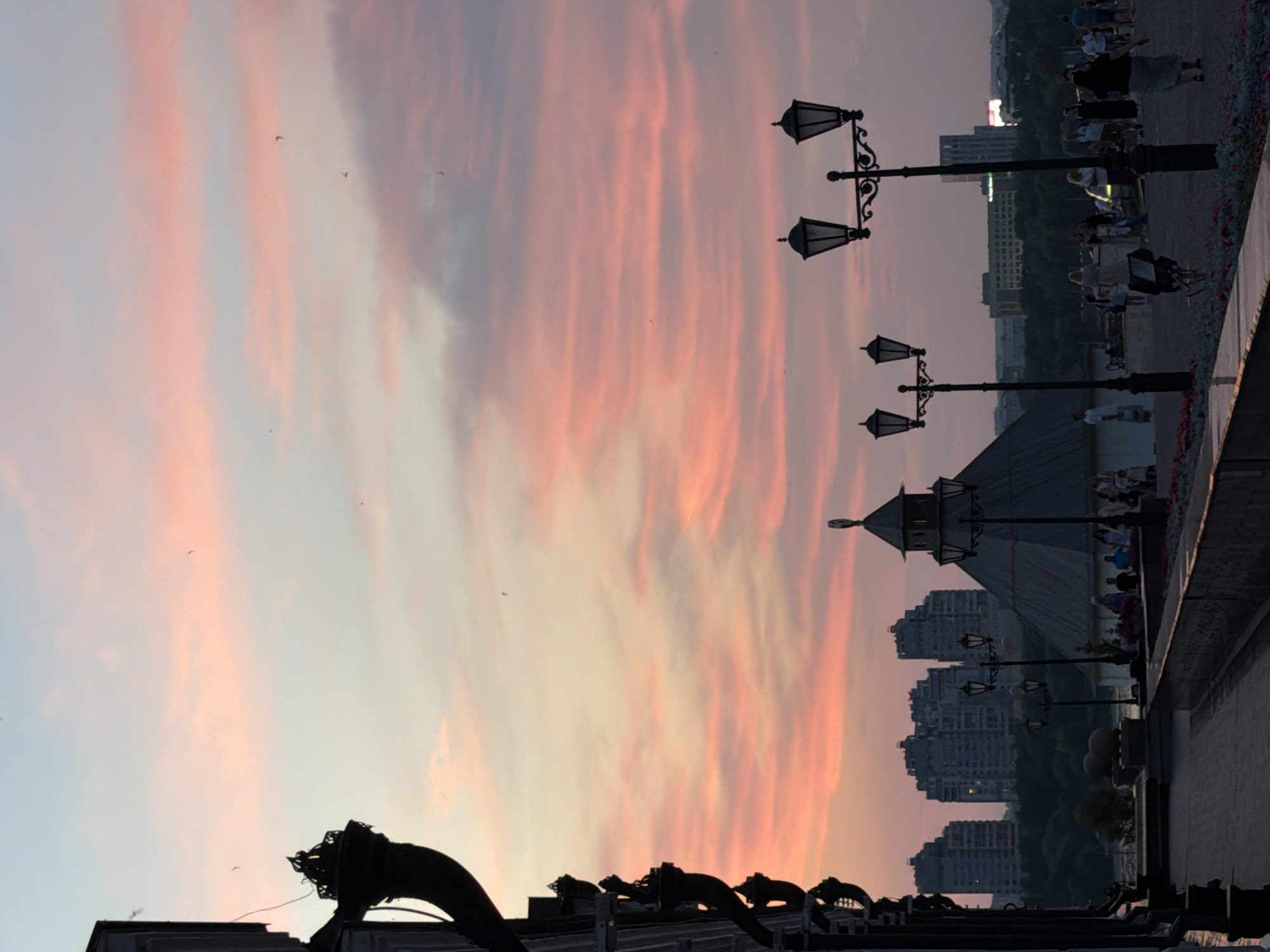 An outdoor shot depicts a city skyline during sunset. The sky is filled with streaks of pink and orange clouds. In the foreground, a silhouette of a building with ornate architectural details is visible on the left. Several lampposts with multiple lights are spaced along a walkway. In the middle ground, a conical-shaped building with a lantern on top is prominent. Behind it, a cluster of modern, high-rise residential buildings stands out against the horizon. Trees are visible between the conical building and the high-rises. A pathway in the foreground is lined with flowerbeds. People are walking along the pathway and gathering near the buildings.
