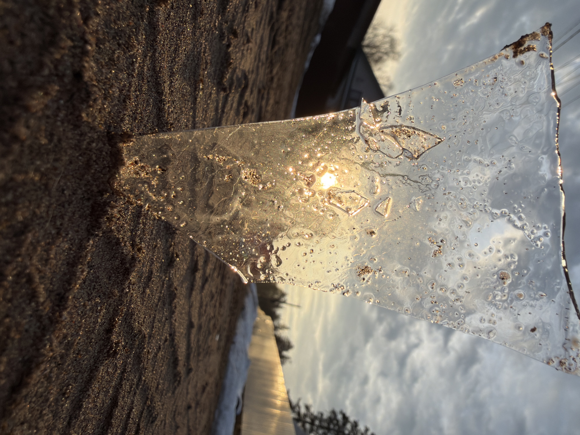 The image shows a triangular piece of ice resting on a sandy or earthy surface, possibly a beach. The sun shines through the ice, creating a bright, shimmering effect. The background reveals a cloudy sky and what appears to be a body of water. Trees and structures are visible in the distance. Without further information or details it is not possible to confirm the location as Chaikovsky, Russia.