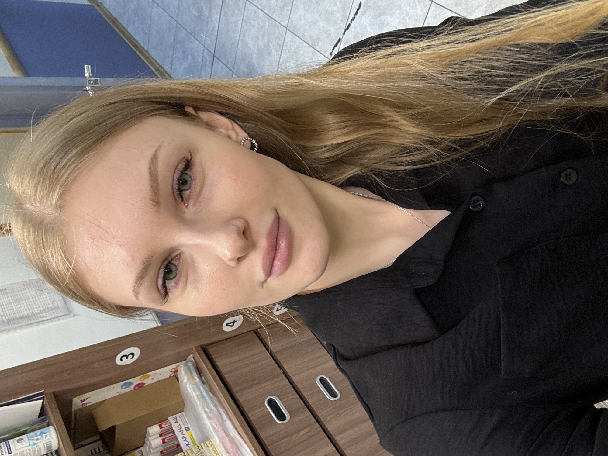 A close-up portrait of a fair-skinned young woman with long blonde hair wearing a black shirt. She is standing in front of a brown wooden cabinet with numbered drawers. The photo was taken in a room. The location is likely in a medical or dental clinic, given the presence of supplies in the open drawer labeled "3". There is no evidence to support the claim that the photo was taken in the city of Perm, Russia.