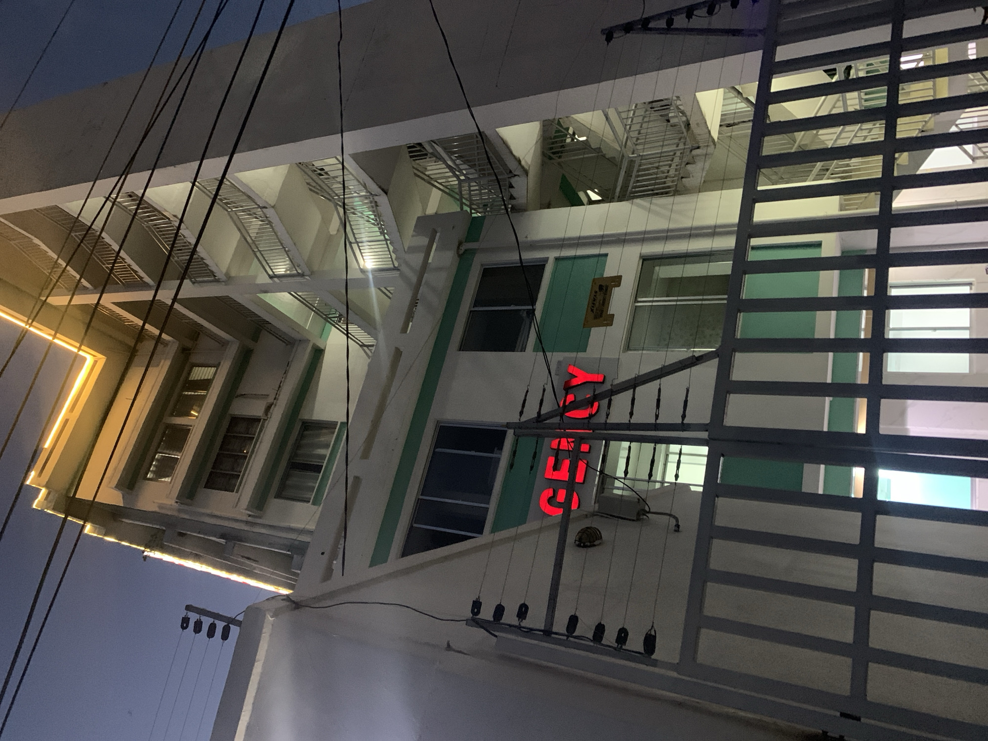 The image shows a building in Dar es-Salaam, Tanzania, identified by a vertically-oriented red neon sign reading "GENCY". The building is light-colored with windows framed in teal. There are metal staircases on the left side of the building. A metal gate obstructs the right side of the image. Power lines are visible crisscrossing the view. The sky is dark, suggesting a night or dusk setting.