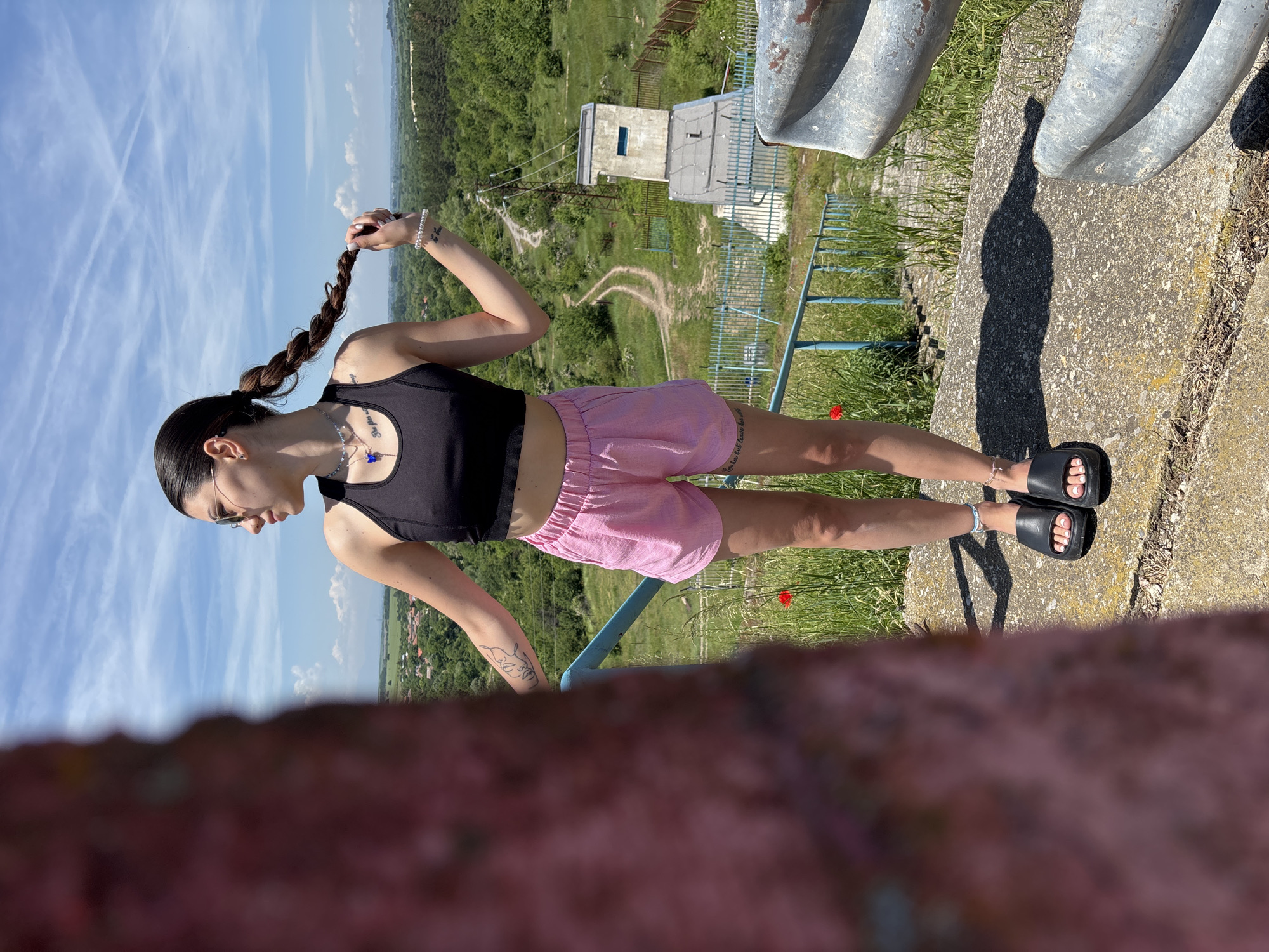 An outdoor, full-body portrait captures a young woman standing on a concrete structure, overlooking a vast, green landscape under a bright, clear blue sky. The image is taken on a sunny day, indicated by the strong sunlight and distinct shadows, suggesting mid-morning or late afternoon.
The primary subject, a woman with dark hair styled in a long braid, is seen from the side, looking towards the distant horizon. She wears a black sports bra or crop top, pink shorts, and black slide sandals with white pedicured toes. Her left hand is casually holding her braid. Notable accessories include a necklace with a blue charm, bracelets on both wrists, an anklet, and sunglasses. Tattoos are visible on her left forearm, depicting an abstract line art face, and on her right wrist, which reads "she goes out". Another bracelet on her left wrist has partially visible text, possibly "she loves".
She is positioned on a rugged concrete ledge or wall, next to what appear to be old, metallic, pipe-like structures. A long, bottle-shaped shadow is cast on the concrete. Below the ledge, a lush, green, hilly terrain stretches out, dotted with trees and a winding dirt path. In the middle distance, a small, light-colored concrete building, possibly an old watchtower or a utility structure, stands amidst the greenery. A blue metal fence or railing runs along the edge of the concrete structure. In the far background, the landscape continues with distant hills and what might be a glimpse of a village. The sky above is largely clear, featuring several prominent contrails forming an 'X' pattern. A single red poppy flower is visible in the grass near the concrete ledge. The setting, in Ognyanovo, Bulgaria, suggests a scenic rural viewpoint.