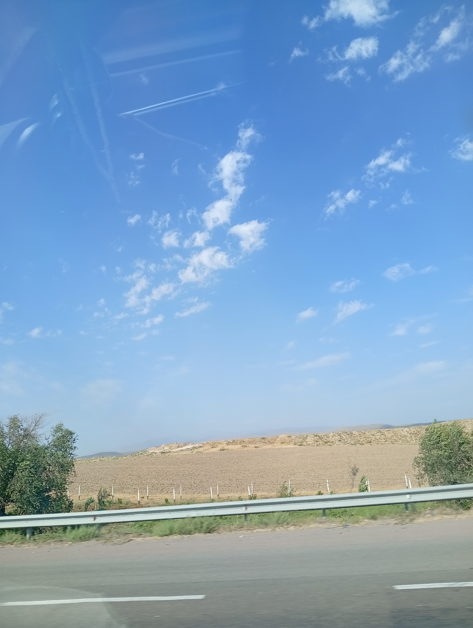 The image depicts a dry, arid landscape with a vast expanse of clear blue sky dotted with scattered white clouds. In the foreground, a gray metal guardrail runs horizontally across the frame, separating a paved road from a barren field. The field itself is characterized by dry, light brown earth and sparse vegetation, with a line of white posts running through it. On the left side of the field, a small cluster of green trees stands out against the otherwise dry terrain. In the distance, rolling hills fade into a hazy horizon. The scene appears to be captured from a moving vehicle, indicated by the slight blur and the reflection of the car's interior on the upper left portion of the sky. The overall impression is one of a rural or semi-rural environment, likely in a region with a warm climate.