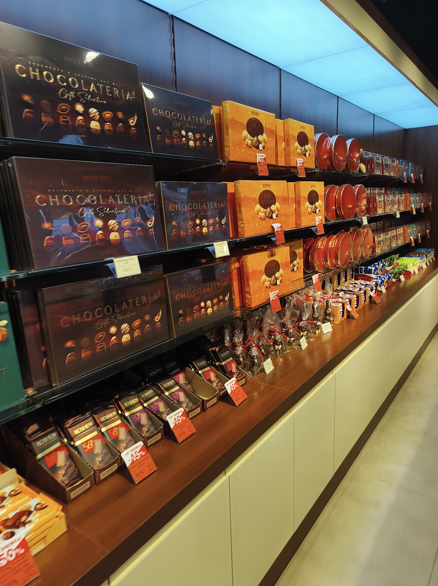 The image depicts a retail display of chocolates in a store, likely in Ternopil, Ukraine, given the Cyrillic text on some packaging. The display features multiple shelves stocked with various chocolate products.
On the top shelf, large, dark-colored boxes labeled "CHOCOLATERIA Gift Selection" are prominent. Next to these, several bright orange boxes from the brand "ROSHE N" are arranged, some with price tags indicating discounts. Further to the right, there are red cylindrical containers and rows of smaller packaged chocolates.
The middle shelf mirrors the top with more "CHOCOLATERIA Gift Selection" boxes and "ROSHE N" orange boxes. Below these, on the wooden surface of a counter, are numerous individually wrapped chocolate items, some in the shape of bunnies and other figurines, many of which are clear-packaged with red ribbons. There are also several rows of smaller chocolate bars displayed in cardboard holders, some of which have discount tags.
The lighting appears to be from above, with a bright, possibly LED, panel casting a cool light. The shelves are made of glass or a similar transparent material, and the display counter is a rich, dark wood. The background shows a wall with wooden paneling and a subtle shelving system. The overall impression is of a well-organized and inviting confectionery display.