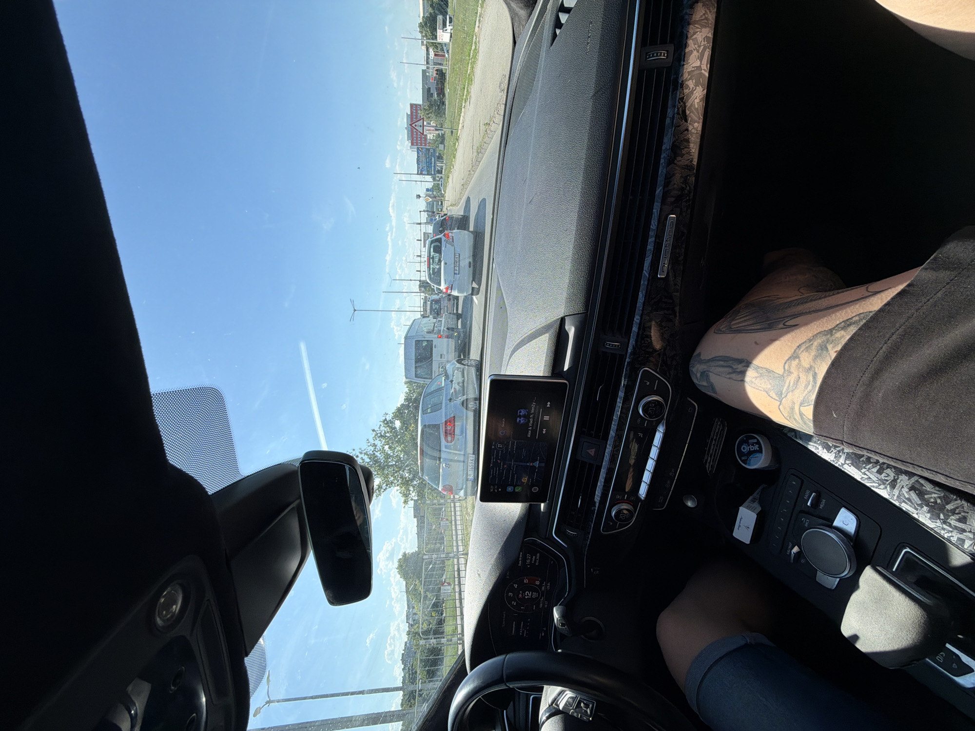The image is taken from inside a car looking forward. The car is stopped in traffic on a sunny day. In the foreground, the dashboard and steering wheel of the car are visible. The driver's left leg, wearing denim shorts, is visible on the left. The passenger's right leg, with tattoos, is visible on the right. The car's infotainment system is displaying a music interface. The road ahead is lined with other vehicles, including a white van, a silver hatchback, and a white sedan. The environment appears to be an urban or suburban area with trees and buildings visible in the background, as well as traffic lights and street signs. The sky is bright blue with some scattered clouds. The license plate of the silver hatchback reads "S 1659 KT". The location is identified as Dolno Ablanovo, Bulgaria.