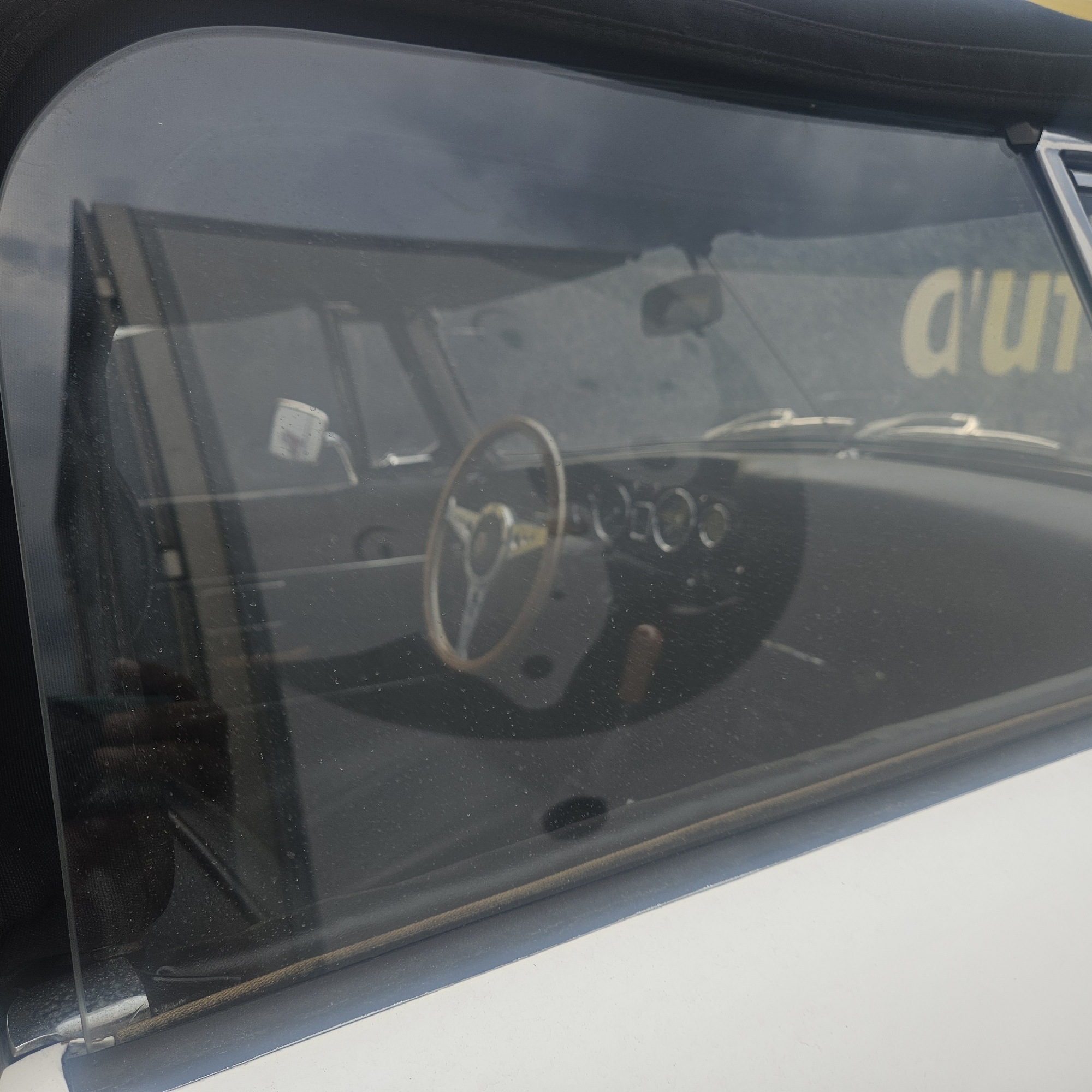 The image shows the interior of a classic car, a convertible, viewed through the side window. The steering wheel, a vintage wooden rim with metal spokes and a central emblem, is prominently displayed. Behind the steering wheel, the dashboard features a cluster of circular gauges and what appears to be a wooden gear shifter. The interior is dark, with reflections on the window obscuring some details. Outside the car, a blurred yellow sign with partial text, "d'un", is visible. The car is likely parked outdoors, as indicated by the lighting and the reflections. The scene is likely in La Jonquera, Spain, given the context. There are no people visible in the image.