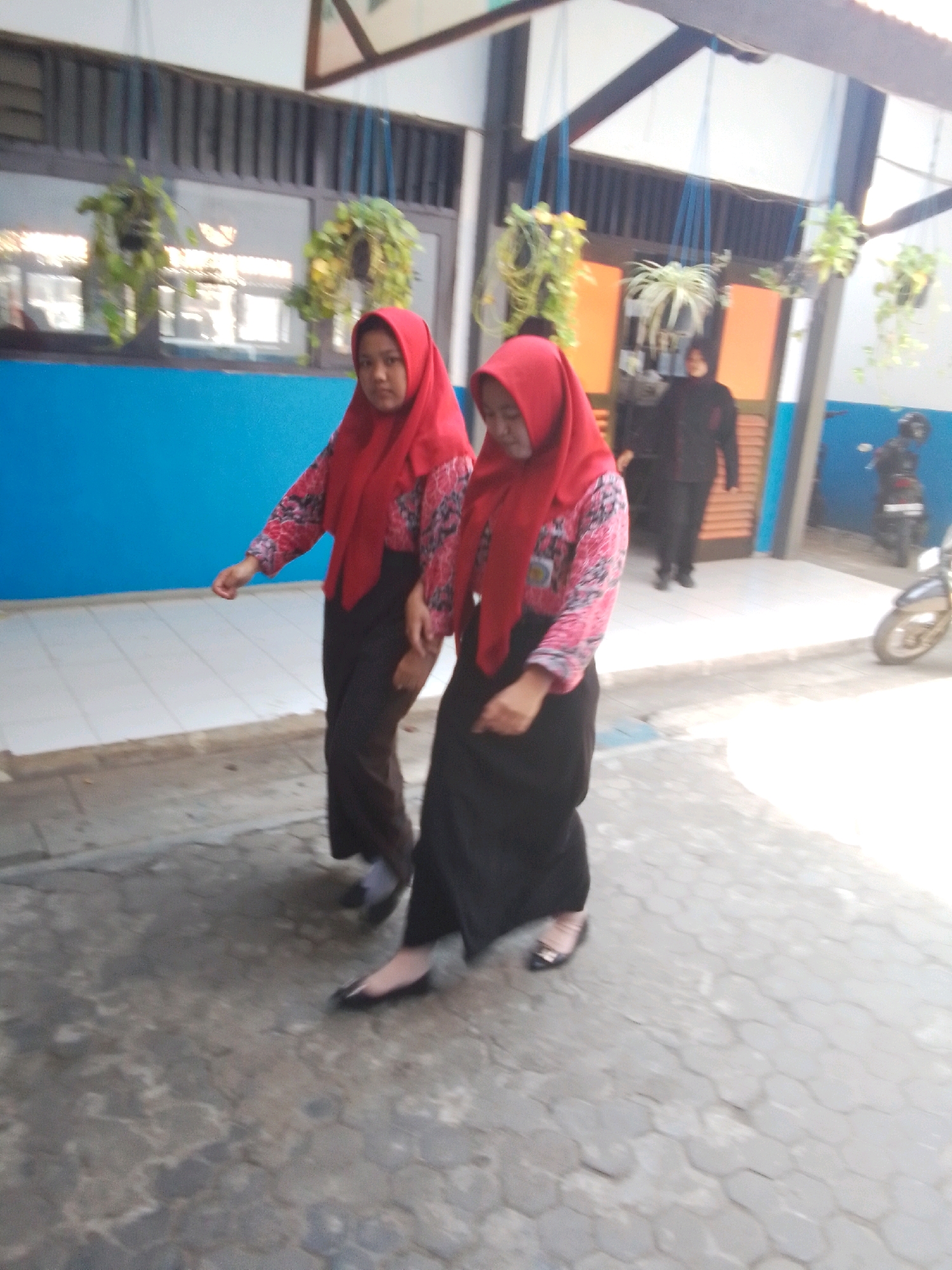 Two young Indonesian females wearing red hijabs and patterned tops over black skirts walk on a paved walkway outside a building with a blue wall. One female on the left is looking down, while the other on the right is looking ahead. Hanging plants are visible in the background, along with a motorcycle and a person in a dark jacket. The scene appears to be in a school or public area in Probolinggo, Indonesia.