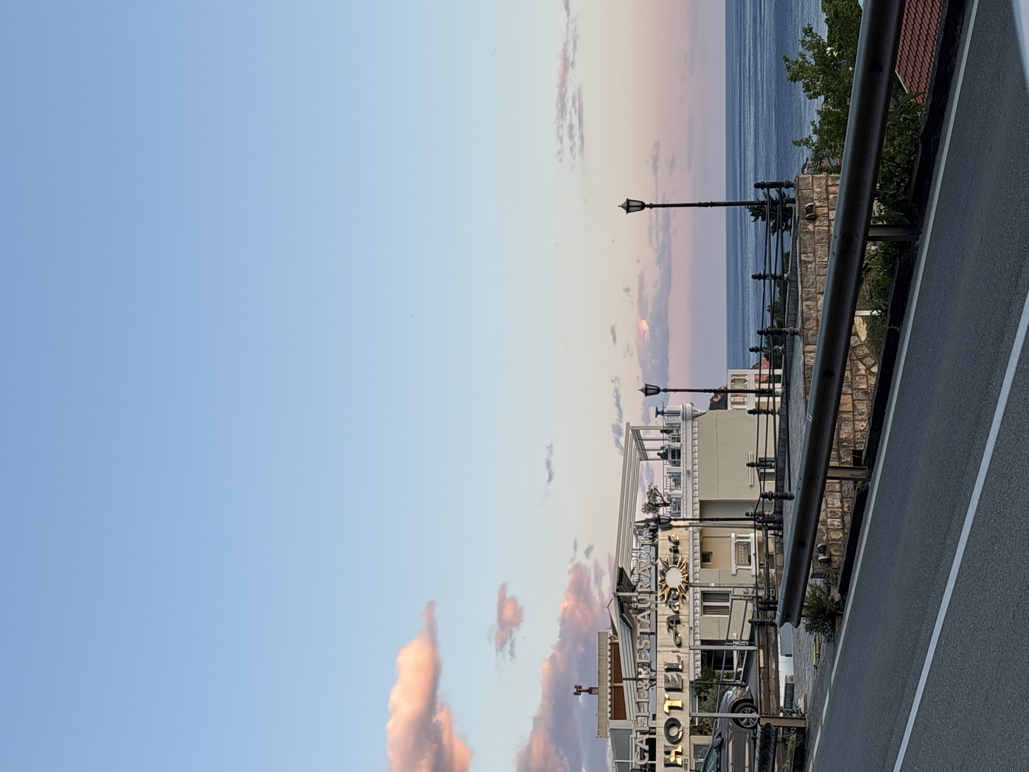 The image shows a street scene in Unknown, Unknown, with a view of a "HOTEL CAFFE & RESTAURANT" building to the right of the frame. A section of a roadside barrier runs along the right edge of the image, parallel to a paved road. Beyond the building, a coastal view includes the sea under a sky with streaks of pink and blue. Street lamps are visible along the roadside. The image is oriented vertically.