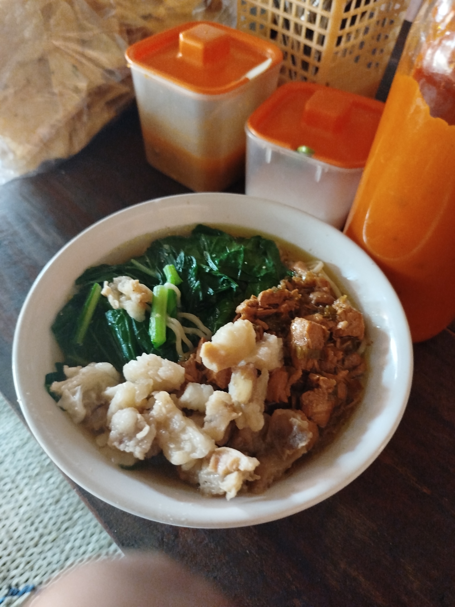 In an indoor setting with a wooden table, a white bowl of food is centered, containing noodles, leafy green vegetables, and pieces of meat. To the right of the bowl, a tall, orange bottle, likely containing a sauce or drink, is visible. Behind the bowl, two small orange-lidded containers and a woven basket are present, along with a bag of food items wrapped in plastic. The scene suggests a meal being prepared or consumed in a casual environment, possibly a home or a small eatery in Pojok, Indonesia.