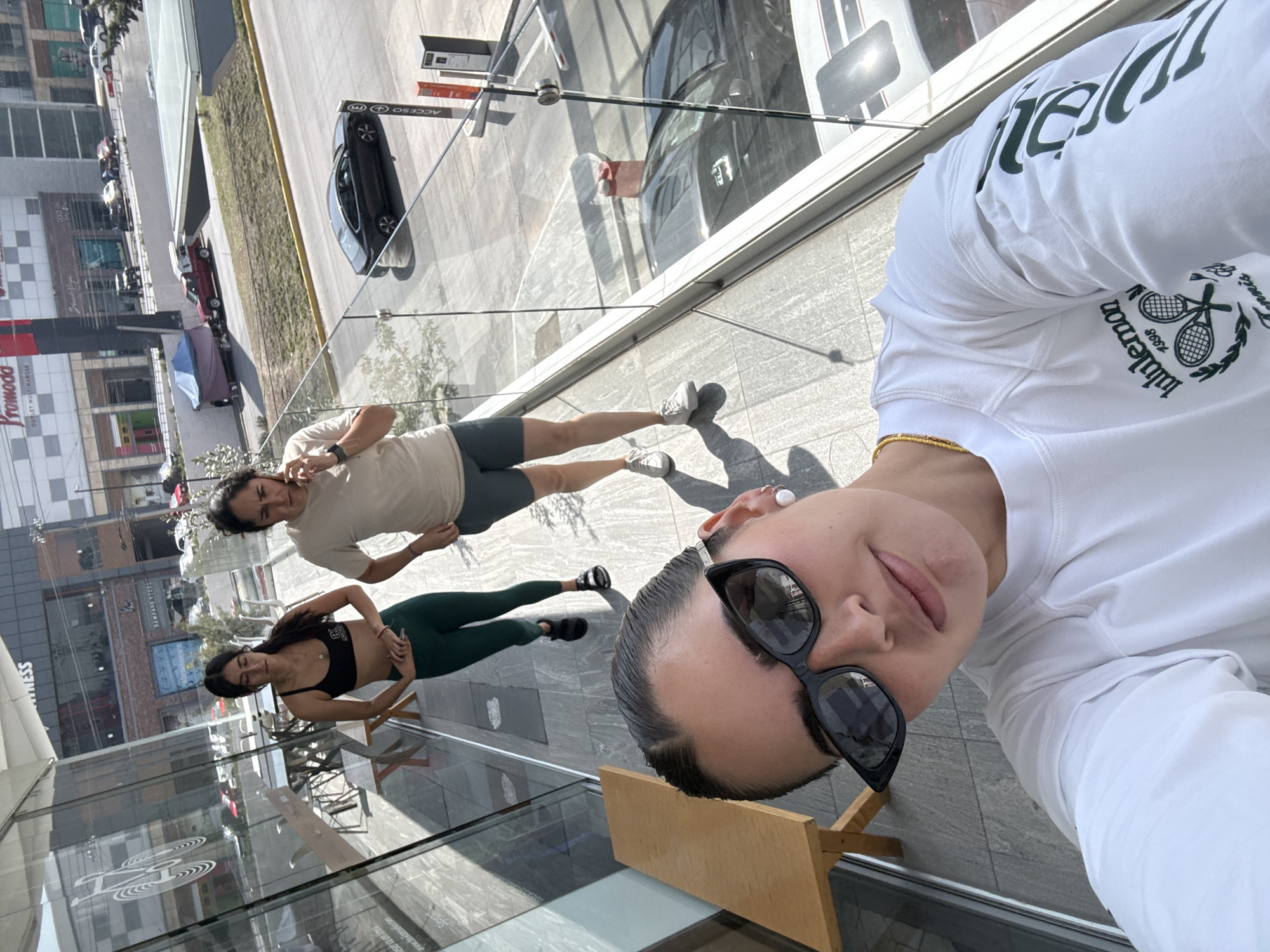 Here is a concise, forensic description of the scene:
The photo depicts three individuals outside a building with a glass facade. The location is Puebla, Mexico, based on the "Promoda Outlet Multimarca" signage visible on the building in the background.
One person, likely the photographer, is positioned close to the camera, wearing sunglasses and a white Lululemon long-sleeved shirt. Further back, two other individuals are standing on the pavement. One wears a beige shirt and grey shorts, talking on their phone. The other individual wears a black sports bra and dark green leggings. The setting appears to be an elevated walkway or balcony overlooking a street with parked cars.