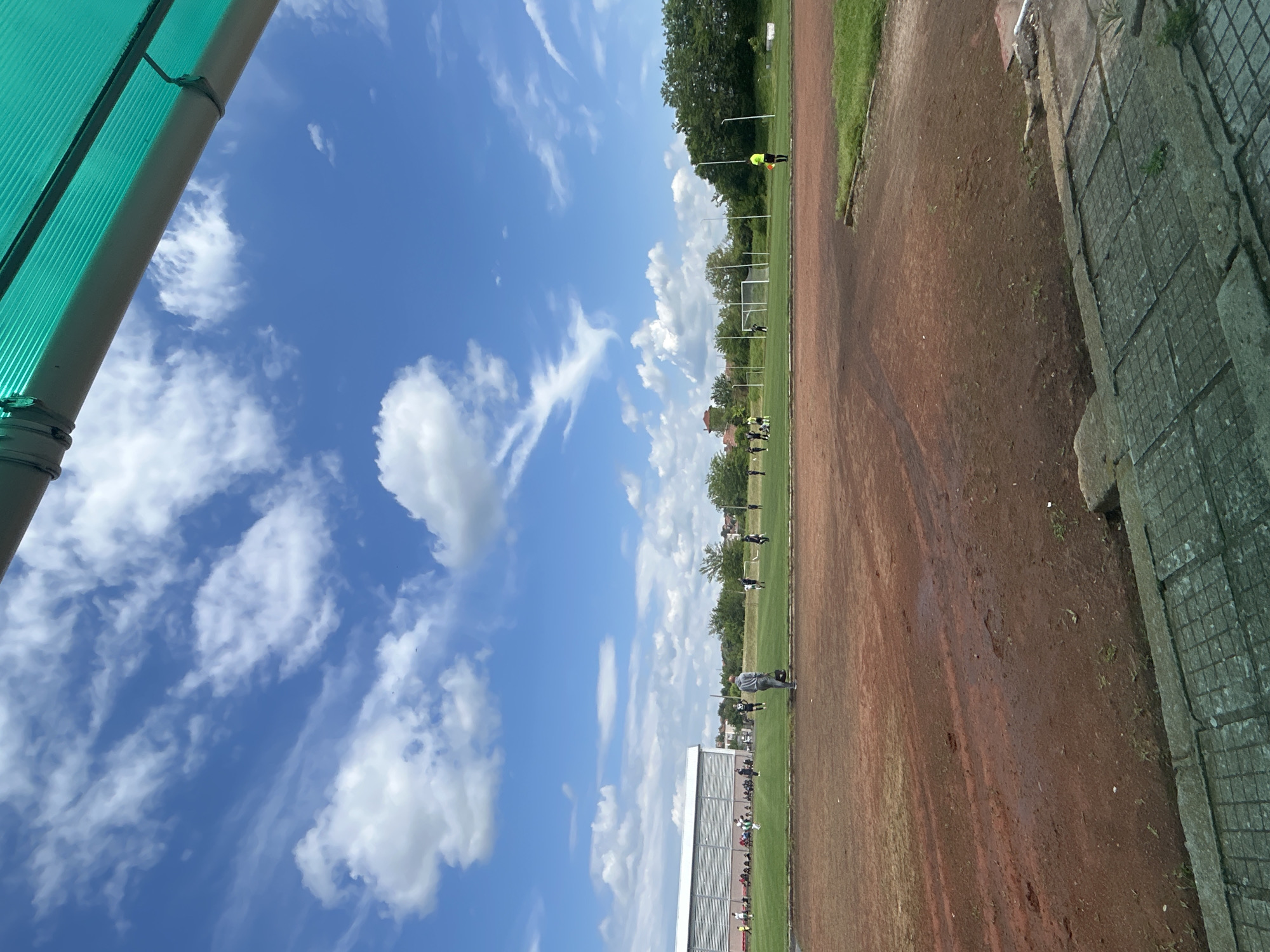 The image shows a soccer field in Unknown, Unknown, with a vibrant blue sky filled with white clouds overhead. On the left side of the frame, there's a section of a green corrugated structure. The soccer field has a reddish-brown track around the perimeter. Several people are on the green grass playing soccer. On the right side of the frame, there's a concrete wall or structure with a grid-like pattern. The scene is bright with natural sunlight.