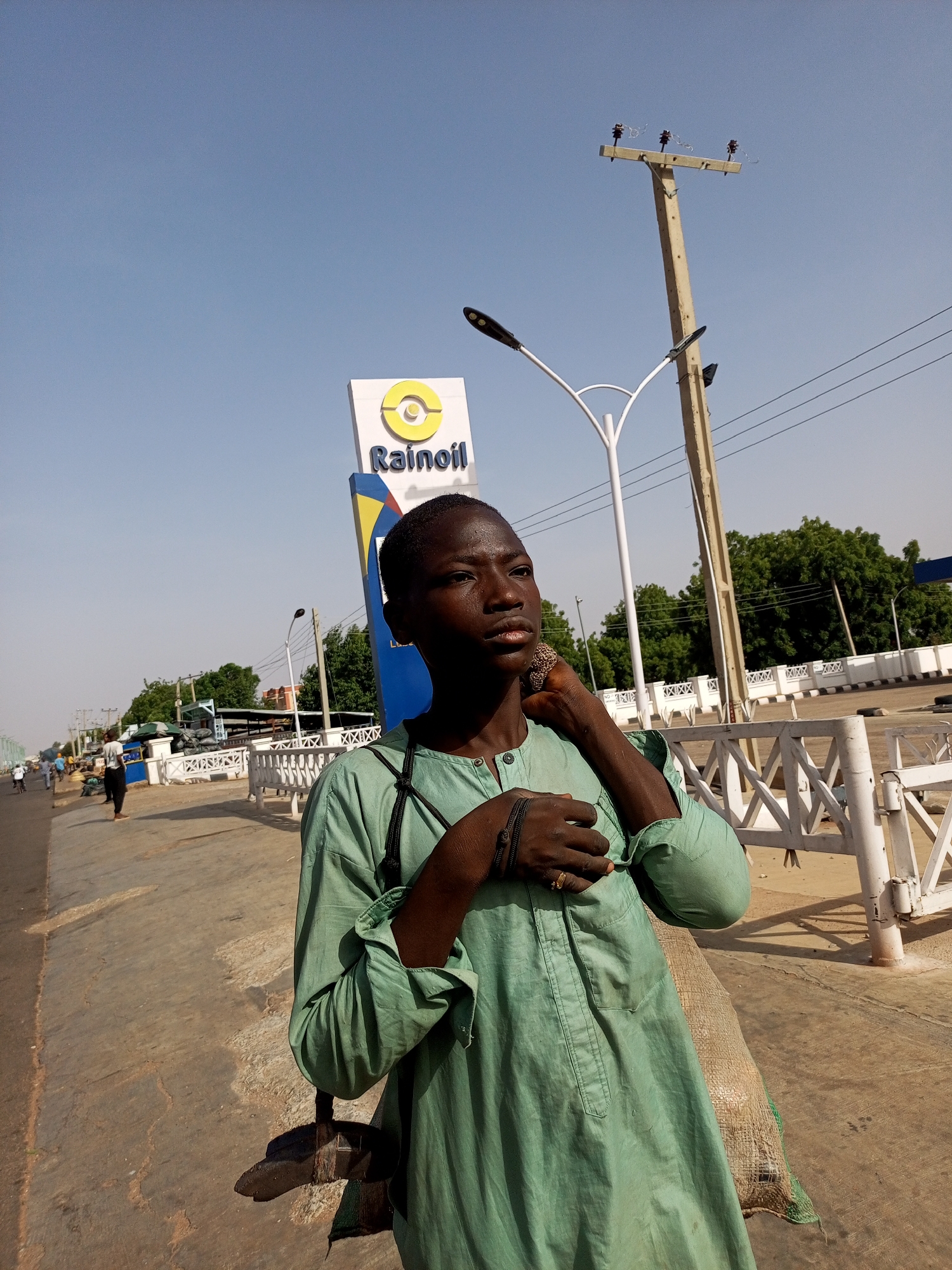 A young, dark-skinned individual stands on a city street in Tempure, Nigeria, next to a Rainoil gas station sign. The person is wearing a light green, long-sleeved garment and has multiple bracelets on their left wrist and a ring on their right hand. A bag rests by their side. Behind them are a white metal barrier, a street lamp, a utility pole, trees, and buildings. Other individuals can be seen in the distance. The sky is bright blue and clear.