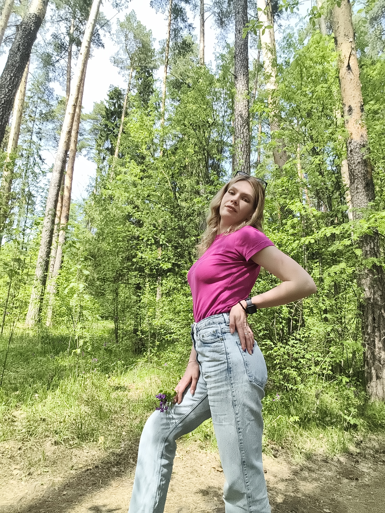 The photograph shows a young woman standing in a forest area. She is wearing a pink t-shirt and light blue jeans. She has blonde hair and is wearing sunglasses on her head. She is holding a small bunch of purple flowers. The background consists of tall trees and lush green vegetation. The location is Izhevsk, Russia.