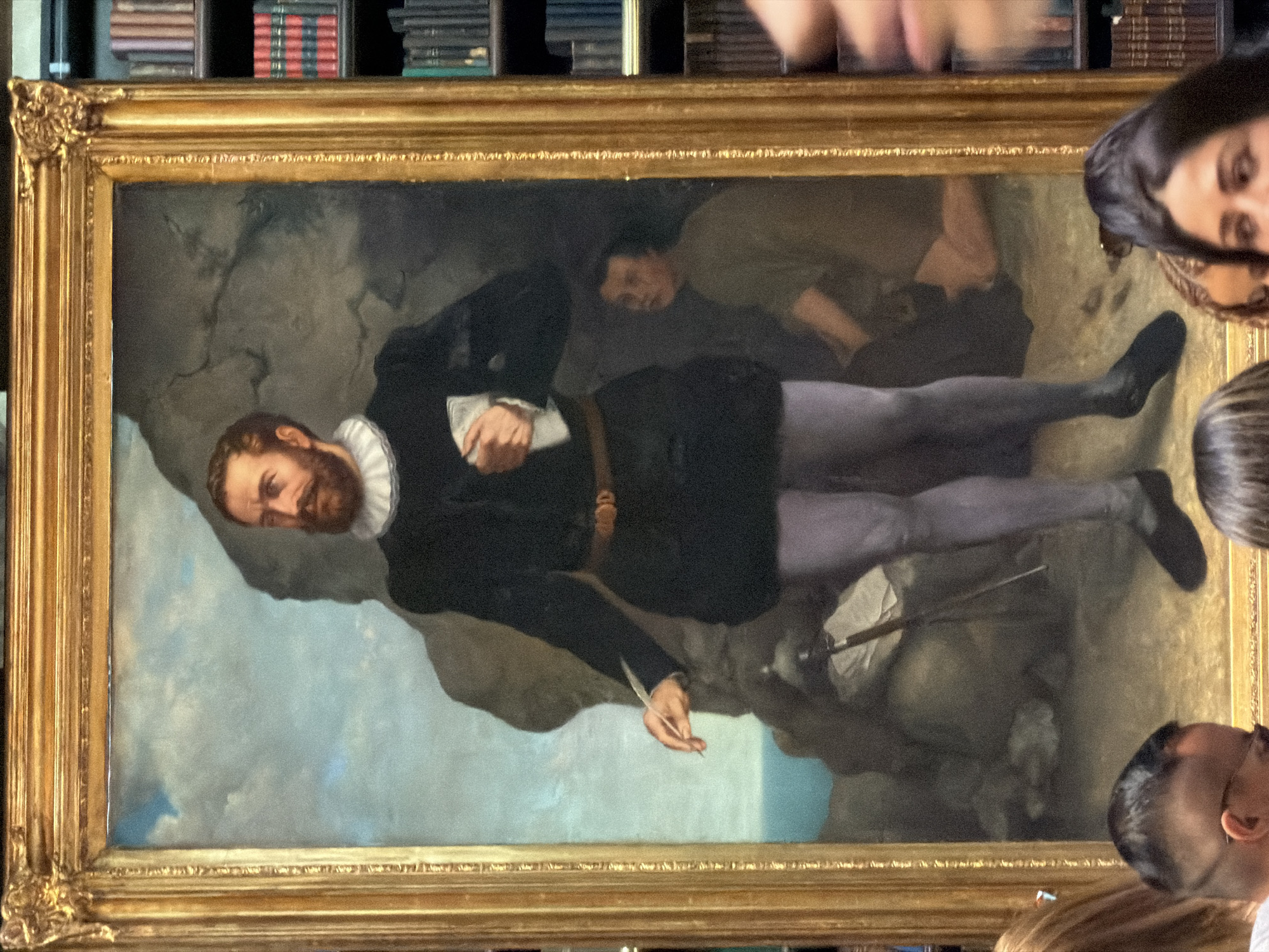 The image shows a framed oil painting of a man in historical attire. The man has a beard and is wearing a dark tunic, a belt with a buckle, and light purple leggings. He is holding a quill pen in his right hand and a piece of paper or cloth in his left hand. Behind him, a figure in a greenish-brown garment is kneeling and looking towards the viewer. The background of the painting depicts a rocky landscape with a cloudy sky and a body of water. The painting is displayed in a ornate golden frame, and is situated in front of bookshelves filled with books. This scene appears to be in a library or museum. The location of this image is not specified as Rio de Janeiro, Brazil.