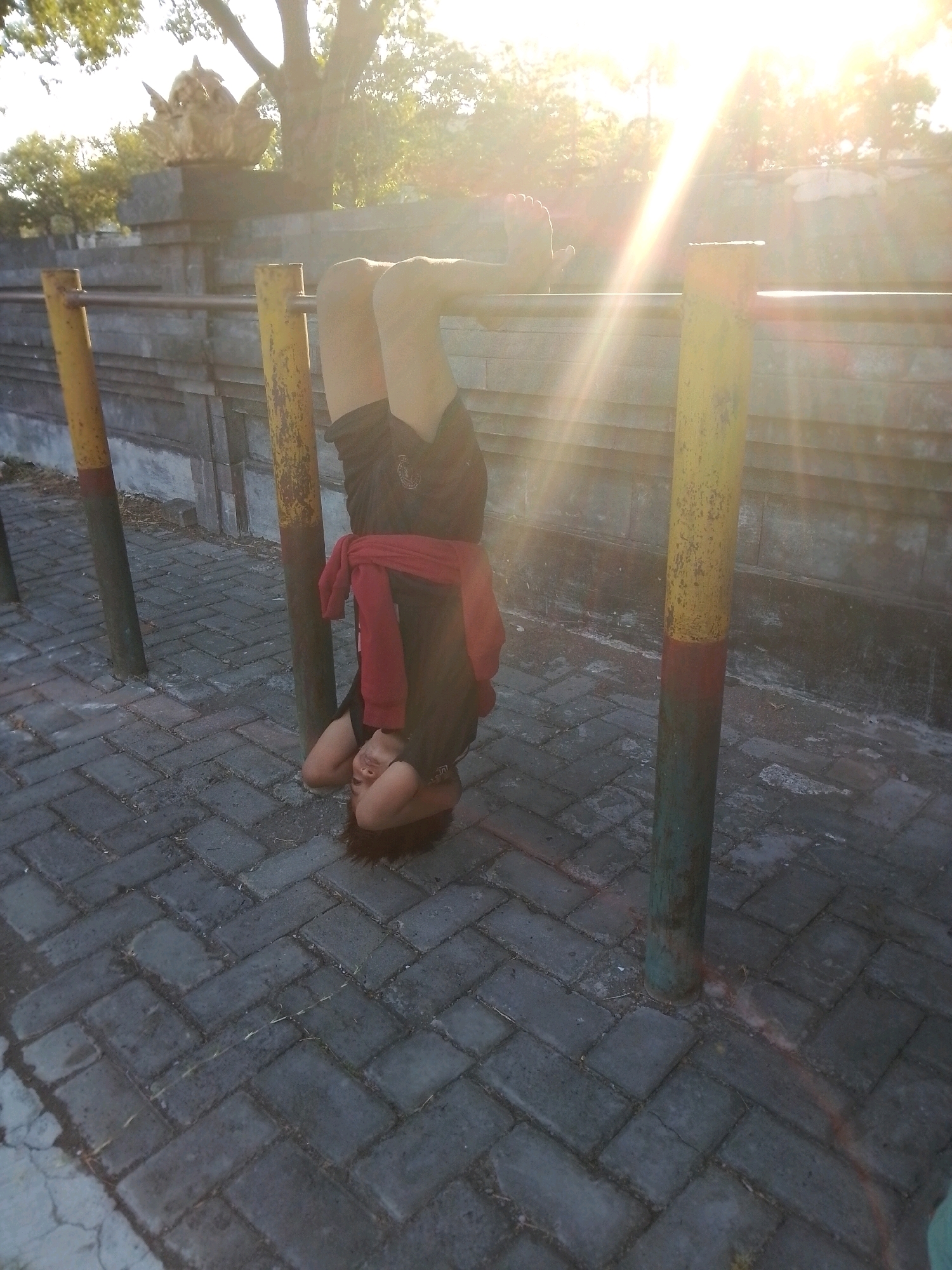 A person is performing a headstand on a horizontal bar at an outdoor gym in Surabaya, Indonesia. The person, wearing a black t-shirt and shorts with a red garment tied around their waist, is suspended upside down with their head resting on the ground. The gym equipment consists of two yellow and green painted poles supporting the horizontal bar. The ground is paved with gray interlocking bricks. In the background, a wall with a golden statue on top is visible, along with trees and a bright sun casting lens flare.