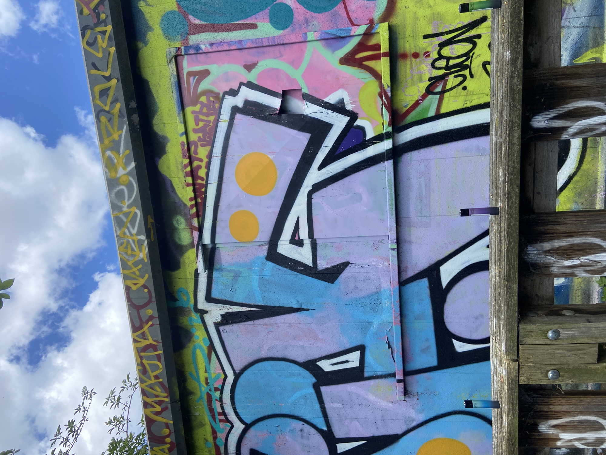 The image shows graffiti on a wall or structure, possibly a bridge or building, under a partly cloudy sky. The graffiti includes several layers of tags, designs, and colors, with some pieces overlaid on others. A wooden structure is visible on the right side of the image, consisting of vertical posts and horizontal beams. The setting appears to be outdoors, urban, and perhaps in a neglected or abandoned area.
The city of London, United Kingdom cannot be confirmed from the image alone.