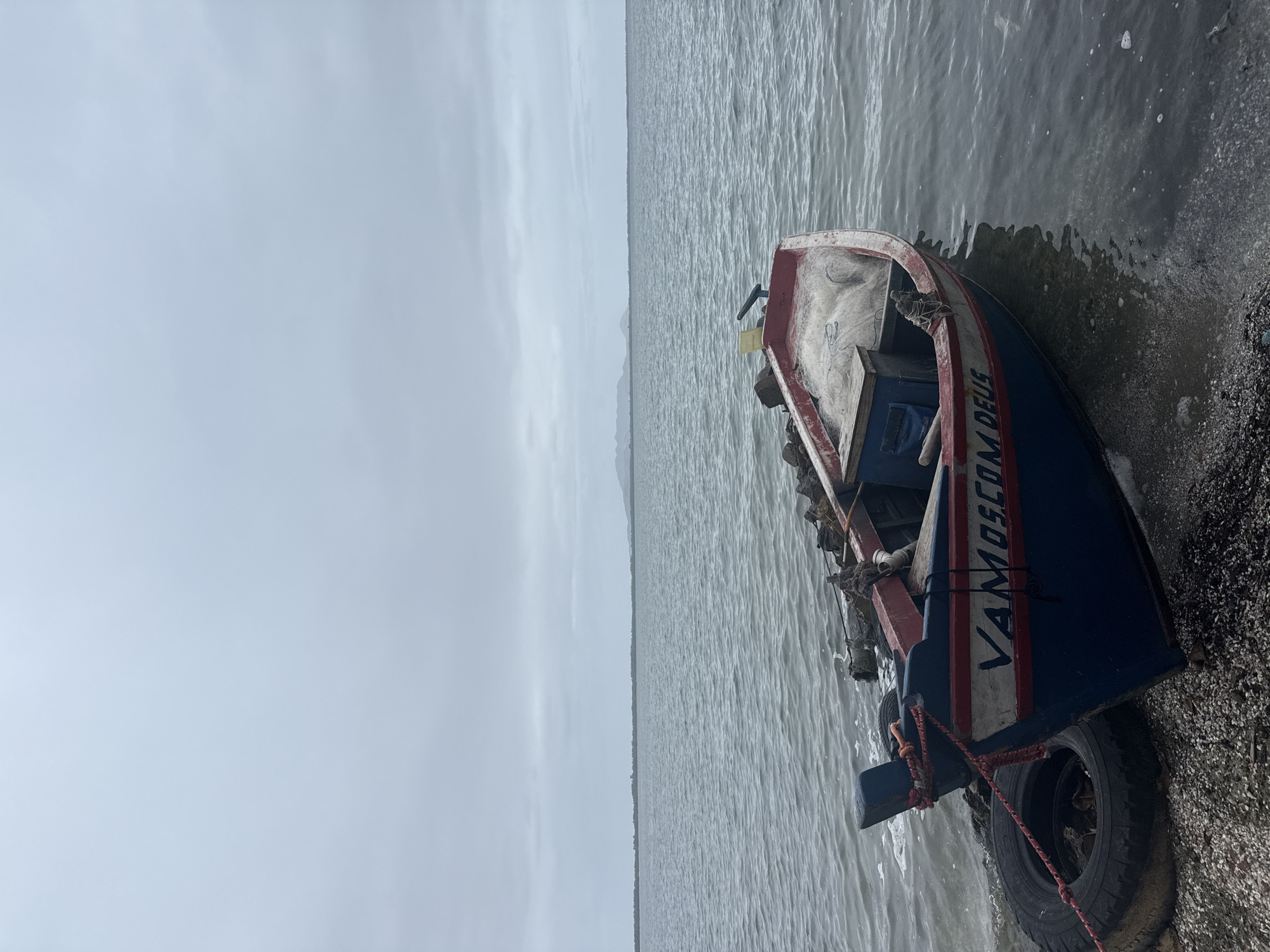 A weathered, blue and red fishing boat is partially beached on a rocky shore under an overcast sky. The boat has the inscription "VAMOS COM DEUS" on its side. A fishing net is visible in the boat, along with a wooden crate. A deflated tire serves as a fender, secured with rope. The calm, gray water of a bay stretches to the horizon, where a faint line of land with distant mountains can be seen. The scene is devoid of human presence and activity. The location is the municipal marina in São Pedro da Aldeia, Brazil.