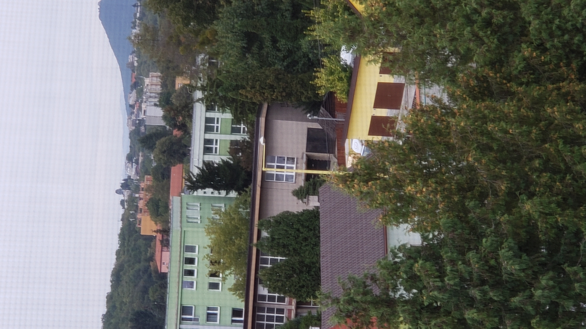 The image shows a panoramic view of a residential area in Prešov, Slovakia, likely from an elevated position. In the foreground, there are several evergreen trees. Behind the trees, there are buildings, including a long, light brown structure with many windows, and further back, a light green building with rows of windows. In the distance, hills covered with trees and some buildings are visible under a cloudy sky. The overall impression is of a suburban or slightly urban environment.