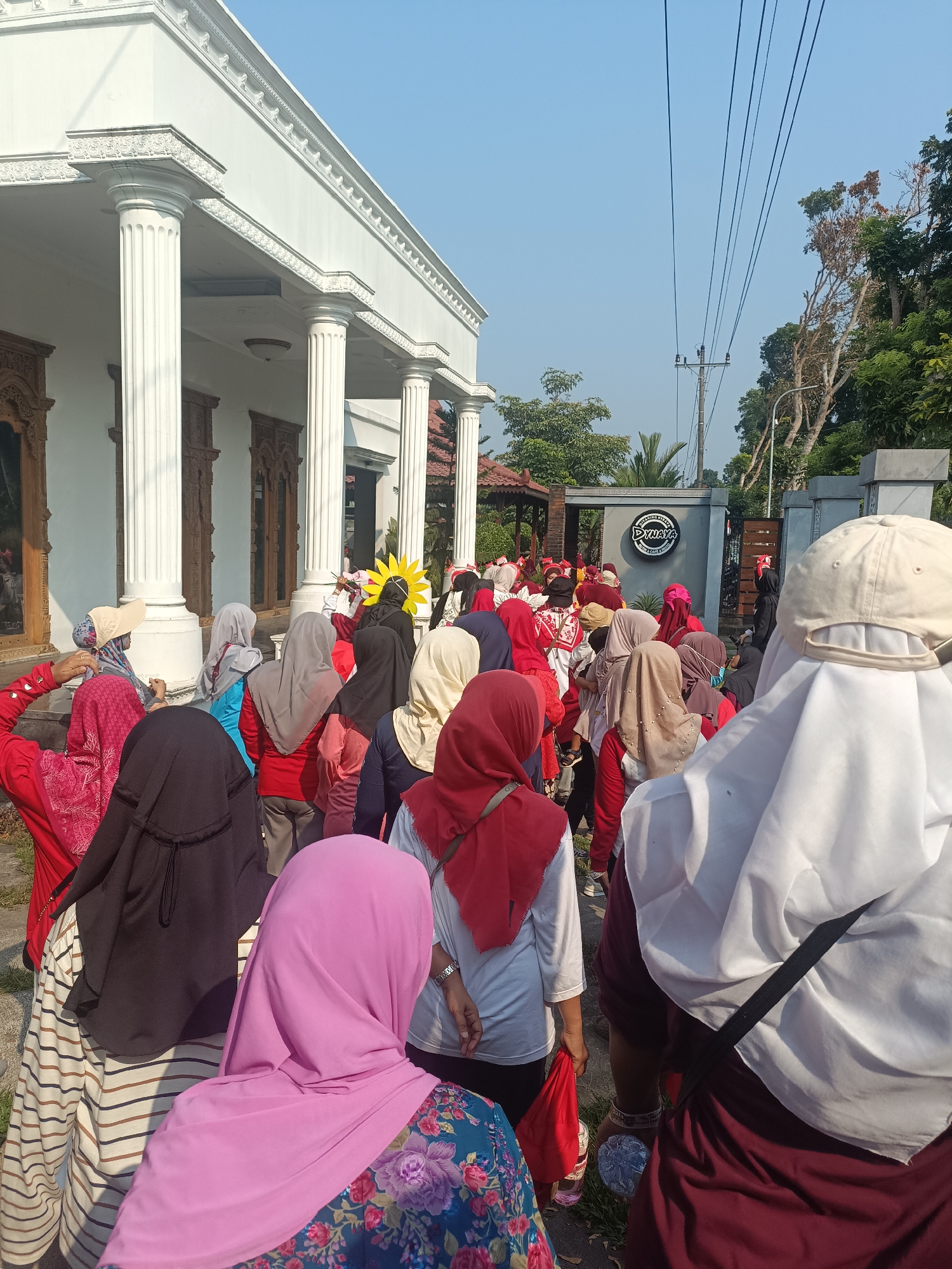 The image shows a group of people, mostly women wearing hijabs, gathered outdoors in what appears to be a residential or commercial area in Sleman, Indonesia. The group is positioned in front of a building with white columns and ornate wooden doors, suggesting a traditional or upscale architectural style. A sign with the text "DINAYA" is visible on a wall in the background. The sky is clear and blue, indicating a sunny day. Some individuals are holding bright yellow flowers, and there is a general sense of gathering or movement among the people. The ground is paved, with some grass and foliage visible. The composition of the image, with its focus on the backs of many individuals, suggests a candid moment or a planned event where the focus is on participation rather than individual portraits.
