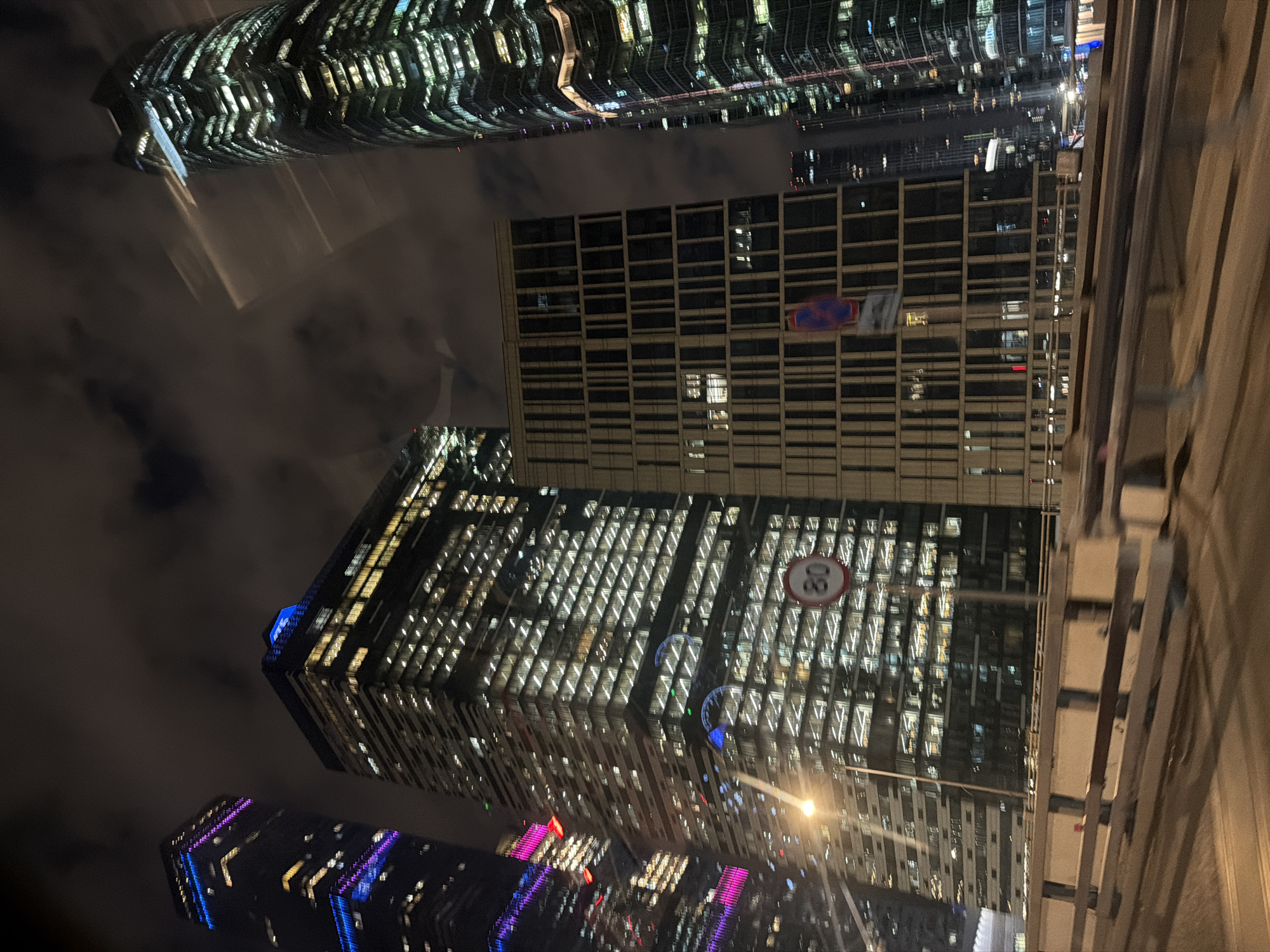 The image shows a night scene in Moscow, Russia, specifically featuring the Moscow International Business Center (Moscow City) skyscrapers. The structures are illuminated with artificial lights, creating a display of geometric patterns and reflections. In the foreground, a building under construction is visible, with vertical wooden beams. The background reveals other skyscrapers with illuminated windows, creating a cityscape against the dark sky. The picture is taken from a low angle.