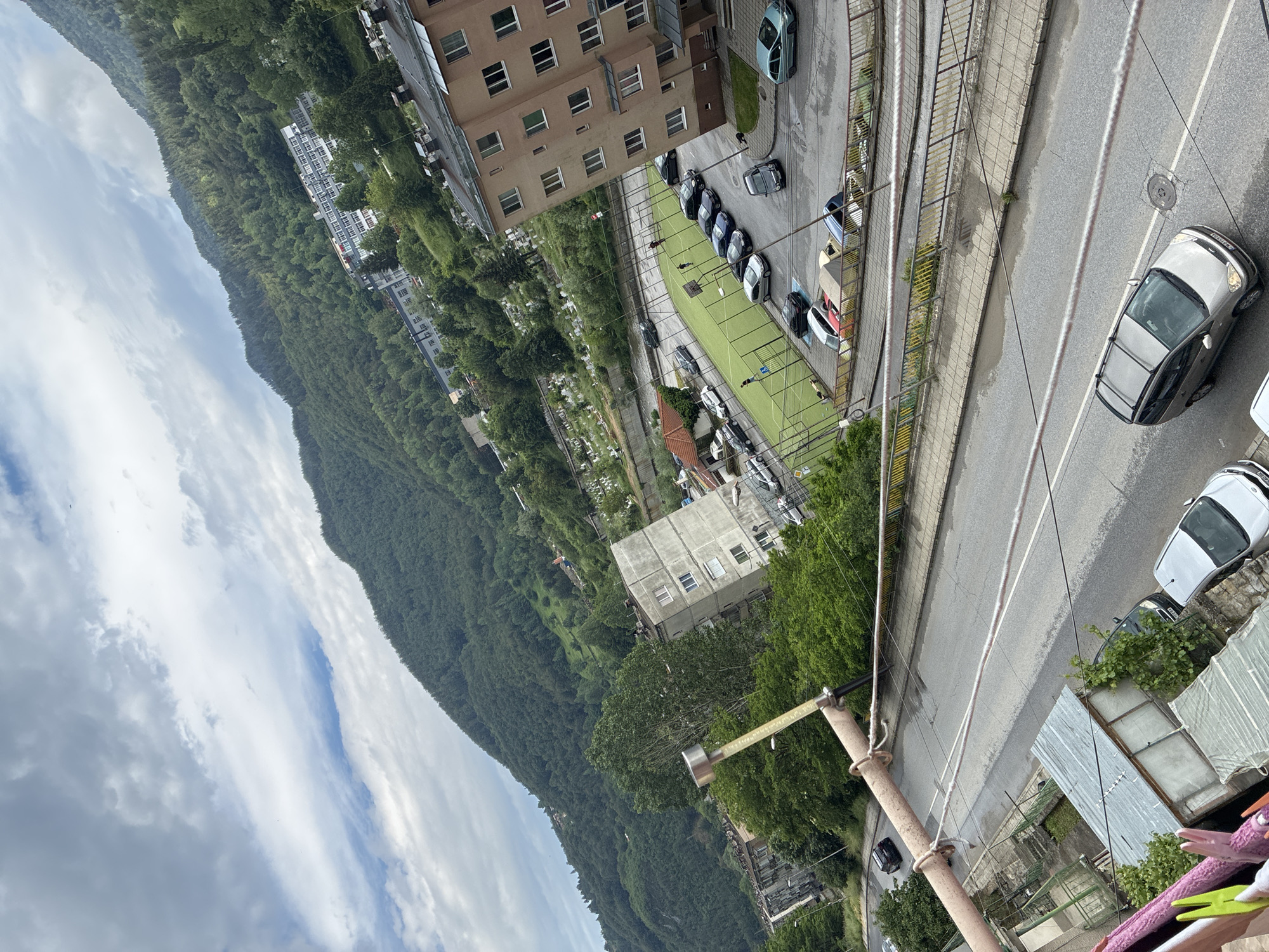 This image captures a diagonal high-angle view of an urban landscape in Vlahovo, Bulgaria, looking over a street and towards forested mountains under a cloudy sky.
The sky occupies the upper left portion, displaying a mix of dense grey and lighter white clouds. The upper right and central background are dominated by a steep, heavily forested mountain range covered in various shades of green deciduous and coniferous trees.
In the mid-ground, below the mountain, several buildings are visible. On the right, a multi-story apartment building with a light brown facade and numerous windows stands next to a parking area. To its left, a green artificial turf sports field with white markings and goalposts is enclosed by a fence. Several parked cars line one side of the field. Beyond the field and above it, a cemetery with numerous light-colored headstones is visible on an incline. Further left, a grey, concrete multi-story building is prominent, with other residential structures scattered along a winding road that parallels the base of the mountain. A larger, multi-story building with a white and grey facade is discernible against the mountain slope in the upper mid-right, possibly an institutional facility.
The foreground features a two-lane asphalt road with a sidewalk on its right. Several vehicles are present on or near the road: a silver minivan/SUV with roof rails is in motion in the lower middle, a silver sedan is partially visible on the far lower left, and multiple other cars are parked or moving in the background and near the sports field. A winding pedestrian path or ramp with yellow railings is visible to the right of the main road, ascending towards the sports field level. From the immediate lower left, a balcony railing extends diagonally, with a metal pole and ropes. A clothesline with colorful clothespins and draped laundry items is visible on the far lower left, indicating the vantage point. Below the balcony, several small, possibly storage sheds with corrugated metal roofs are situated. At least three individuals are visible on the green sports field, appearing to be walking or engaged in activity.