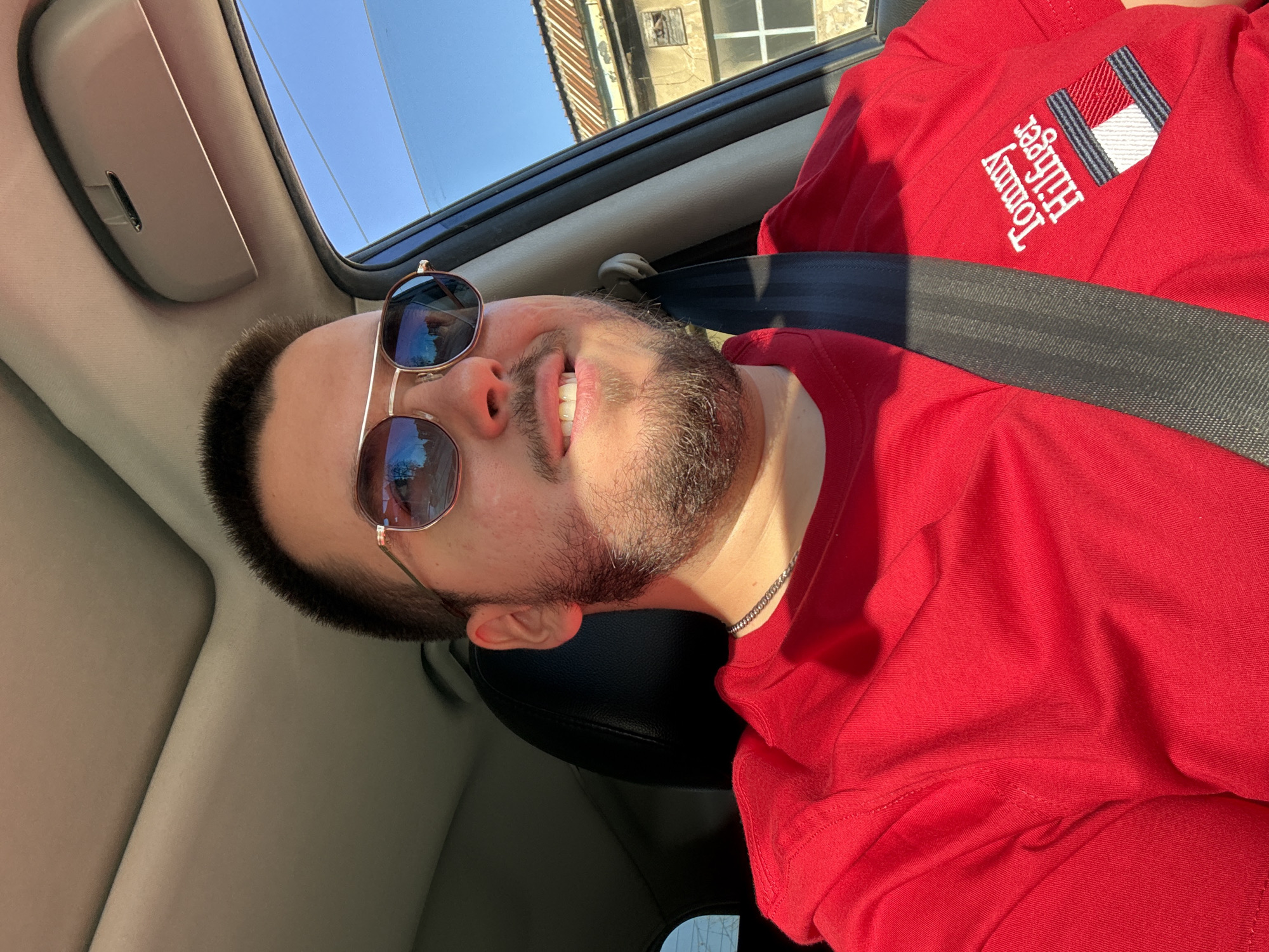 A man is sitting in a car. He is wearing a red Tommy Hilfiger t-shirt and sunglasses. The man is wearing a seat belt. The picture was taken in a car in Sofia, Bulgaria.