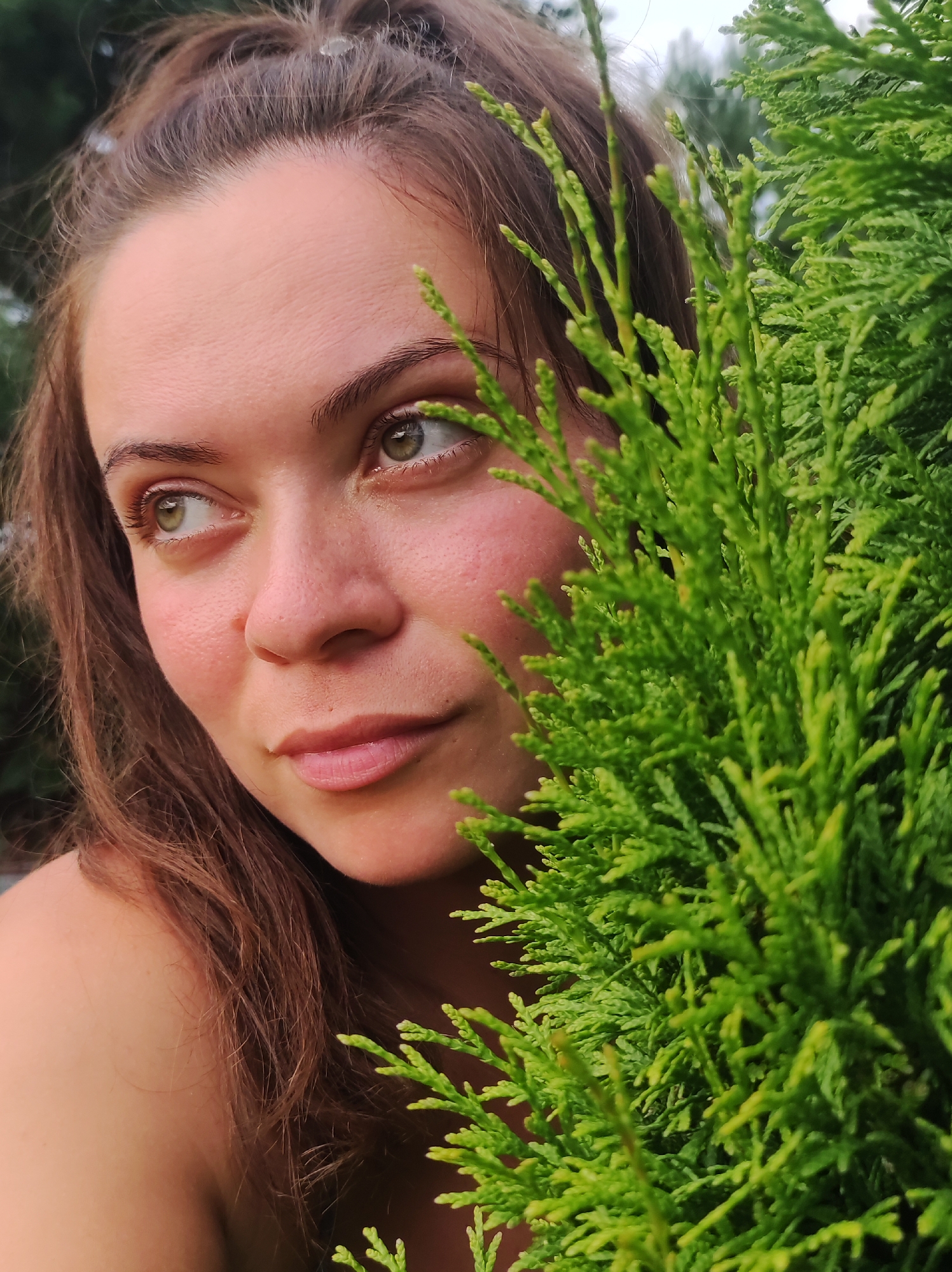 The image is a close-up portrait of a young woman with brown hair and green eyes, partially obscured by lush green foliage. She is looking to her left, with a thoughtful expression. Her skin appears fair, with some rosy blush on her cheeks. The background is blurred, suggesting an outdoor setting, possibly a park or garden. There is no visible indication of the city of Фоки, Russia.