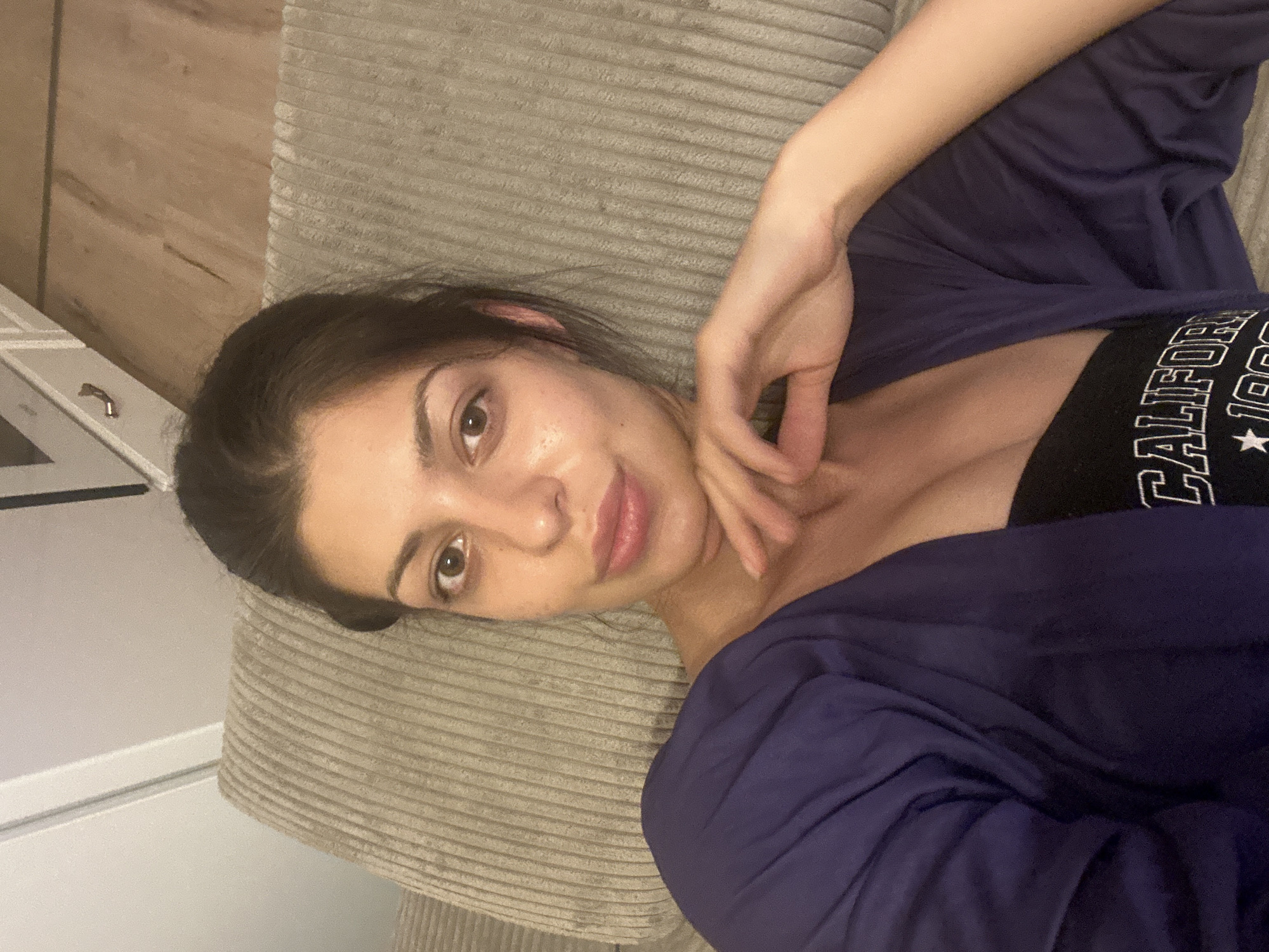 A woman is lying on a ribbed couch, her head resting on her hand. She is wearing a dark purple cardigan over a black tank top with the word "CALIFORNIA" printed on it. The background shows a wooden floor and part of a white kitchen cabinet. The setting is an indoor apartment in Sofia, Bulgaria.