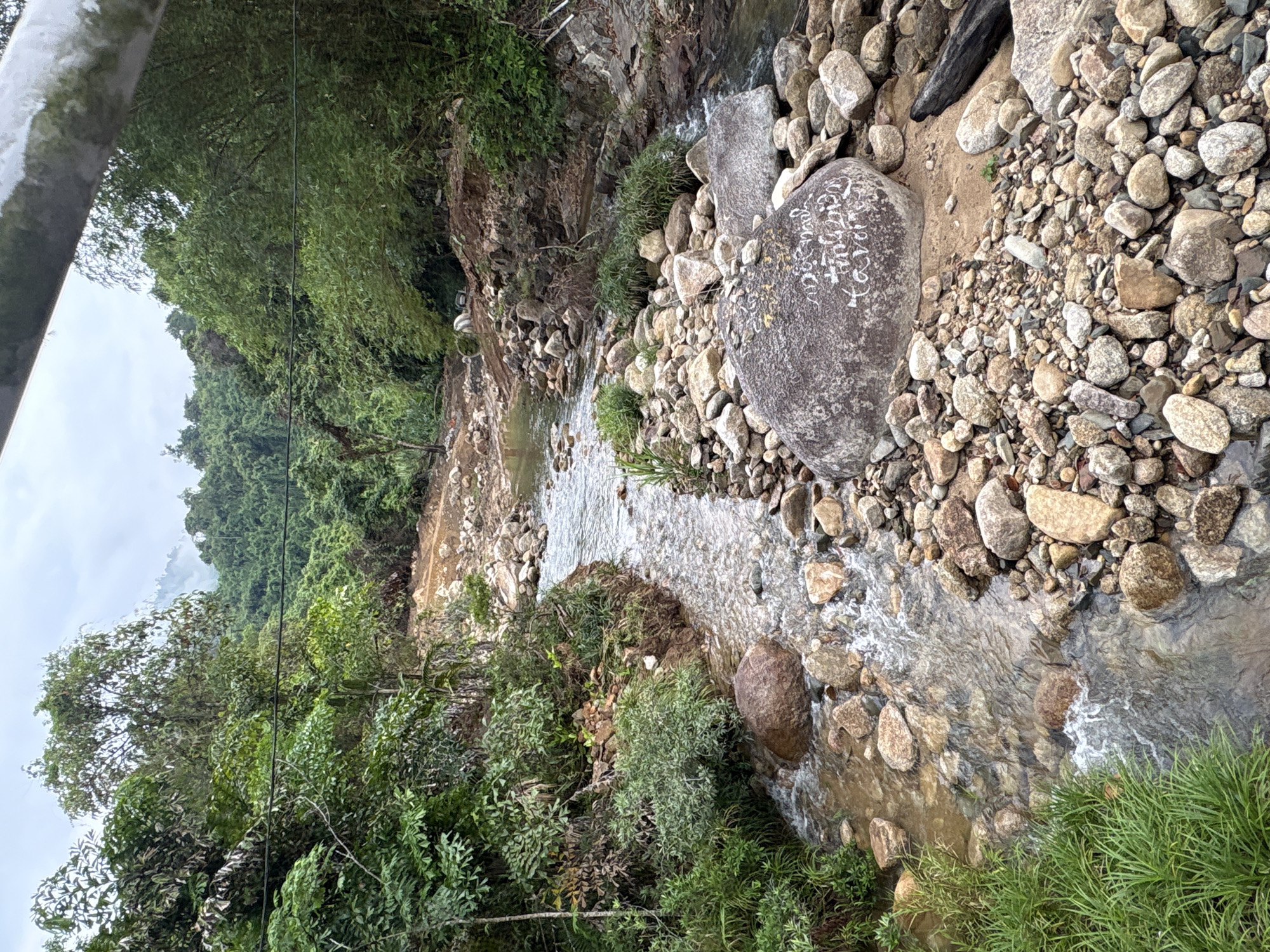 The image shows a stream flowing through a rocky landscape in Khanh Vinh, Vietnam. The stream is surrounded by rocks and vegetation. A large, engraved rock lies near the center of the image. The surrounding area includes trees, shrubs, and smaller rocks. The sky is overcast.
