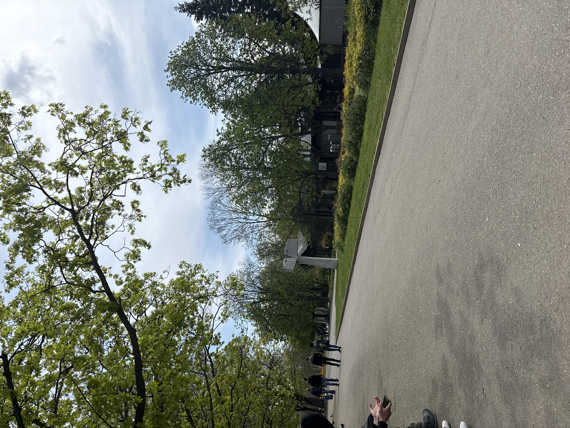 The image shows a paved walkway lined with trees in Sofia, Bulgaria. A group of people is walking along the walkway. A modern stone monument stands in the distance. The sky is partly cloudy.