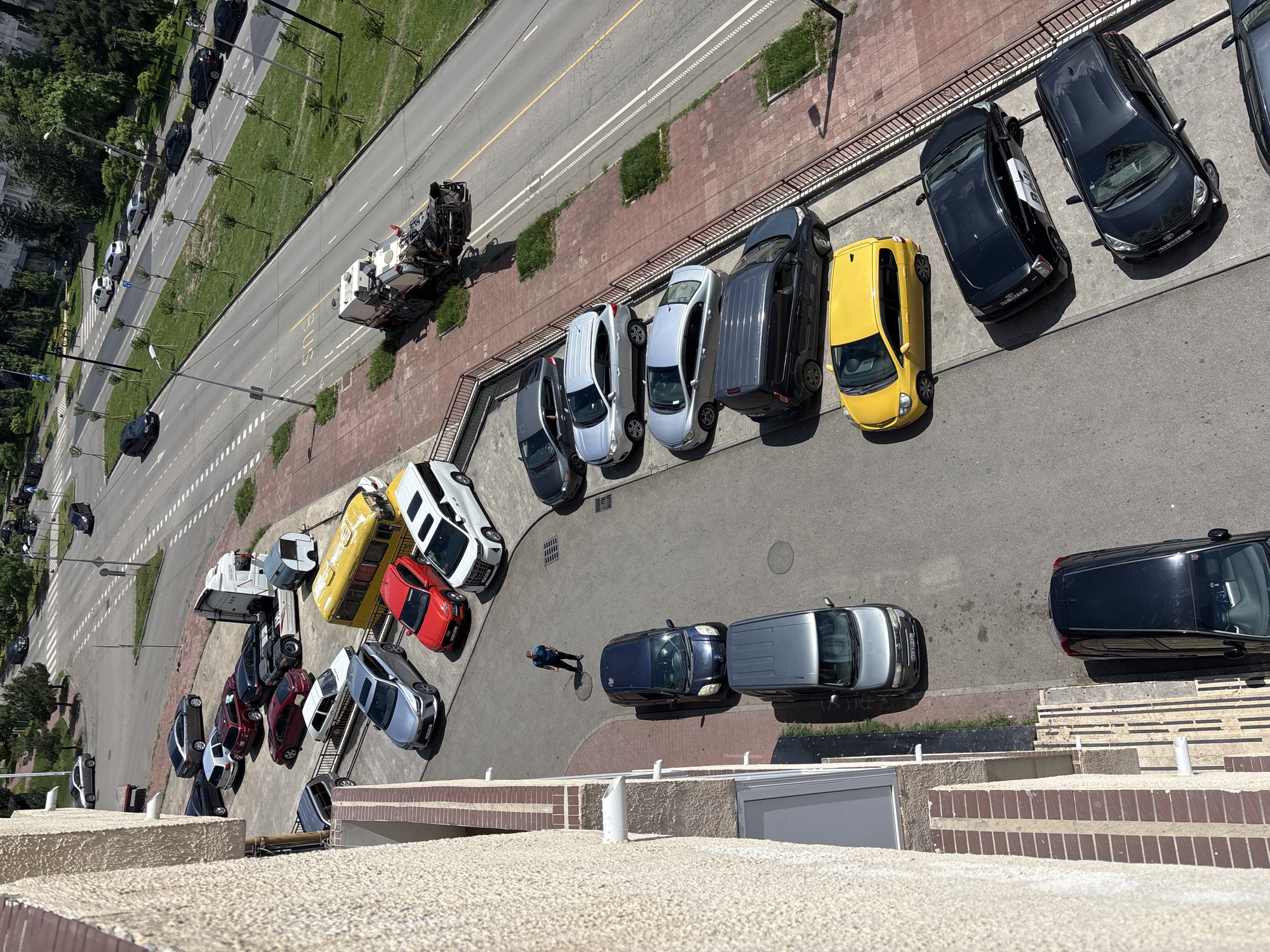 A high-angle photograph shows a street with parked cars, a sidewalk, and a road with moving vehicles, taken from a tall building in Tbilisi, Georgia. The image includes a mix of dark-colored and lighter-colored cars parked along the sidewalk. On the road, a truck and other vehicles are moving. A pedestrian is visible on the sidewalk near parked cars. The building from which the photo is taken has a beige-colored brick exterior.