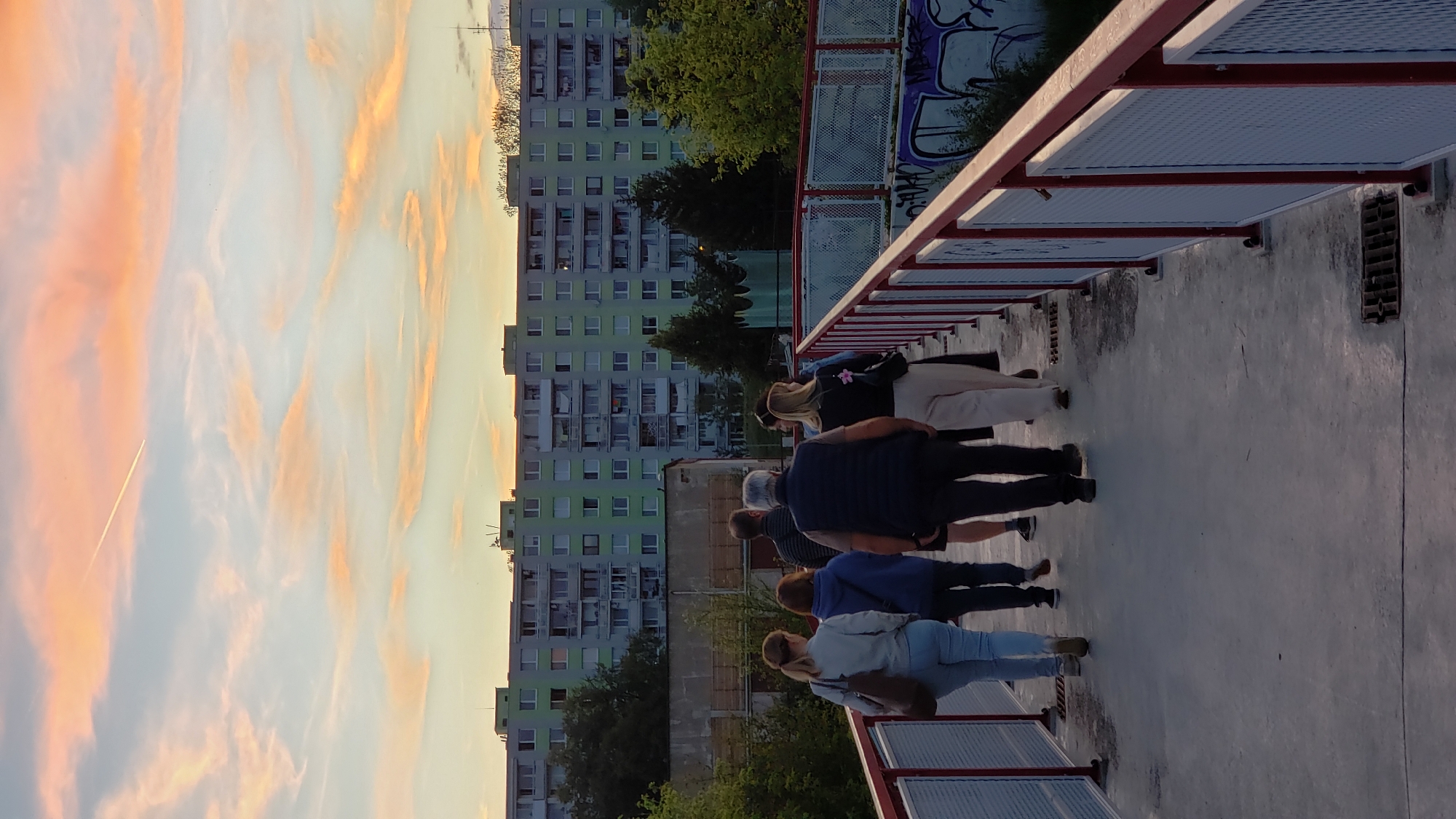 A group of six people are walking away from the camera on a pedestrian bridge. The bridge has a red railing with white mesh inserts. In the background, there is a large, light green apartment building with many windows. Above the building, the sky is a mixture of pale blue and orange clouds, with a contrail from an airplane visible. The ground of the bridge appears to be concrete. The building in the background is located in the Šebastovce neighborhood of Prešov, Slovakia.