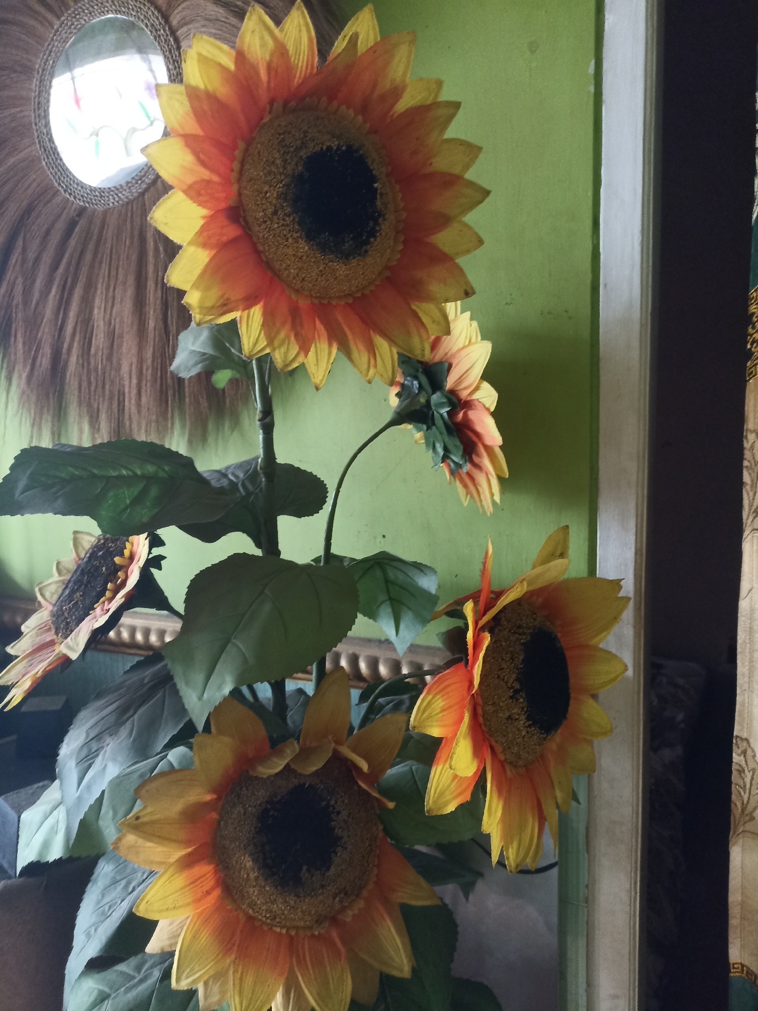 An artificial sunflower arrangement is displayed indoors. The sunflowers have orange and yellow petals with dark centers. Several large green leaves are attached to the stems. In the background, a green wall is visible, along with a mirror framed with natural fibers and a doorway with a patterned curtain. The scene is likely a residential interior in Pandanarum, Indonesia, given the context provided, but the image itself does not contain explicit identifiers of the location.