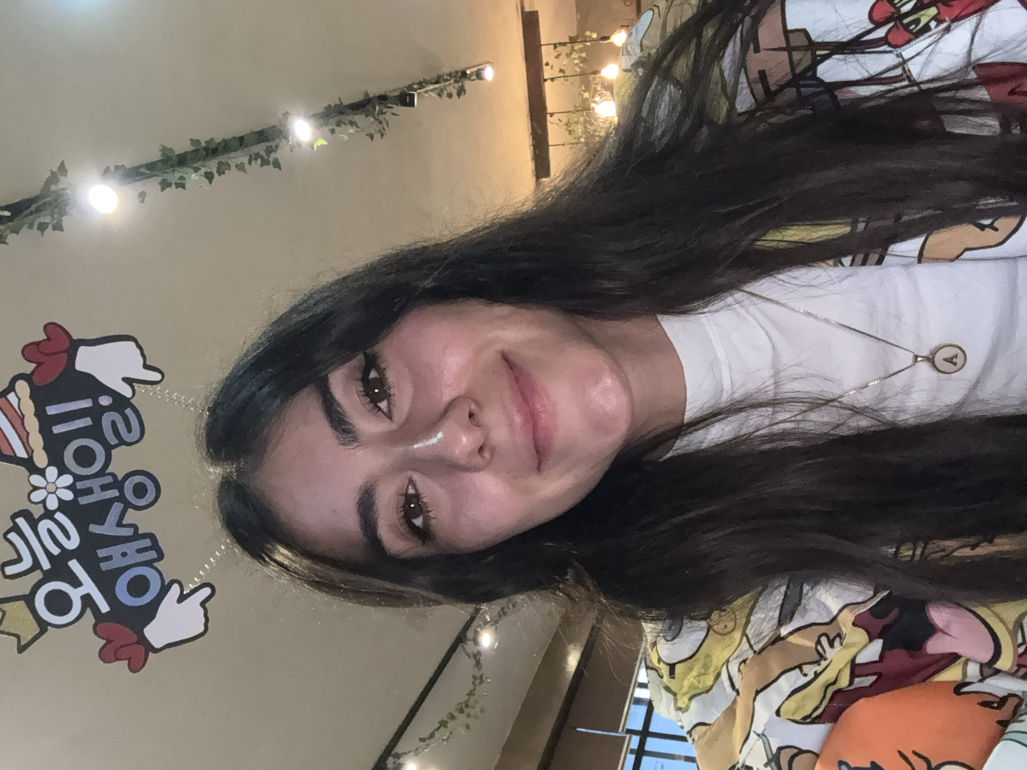 A young woman with long dark hair is looking into the camera with a slight smile. She is wearing a white t-shirt and a necklace with a circular pendant. Behind her is a wall decorated with lights and greenery. To the left, there's a sign with Korean text and cartoon figures. The lighting is warm and artificial. The location appears to be indoors, but the exact location within the city of Puebla, Mexico, cannot be determined from the image alone.