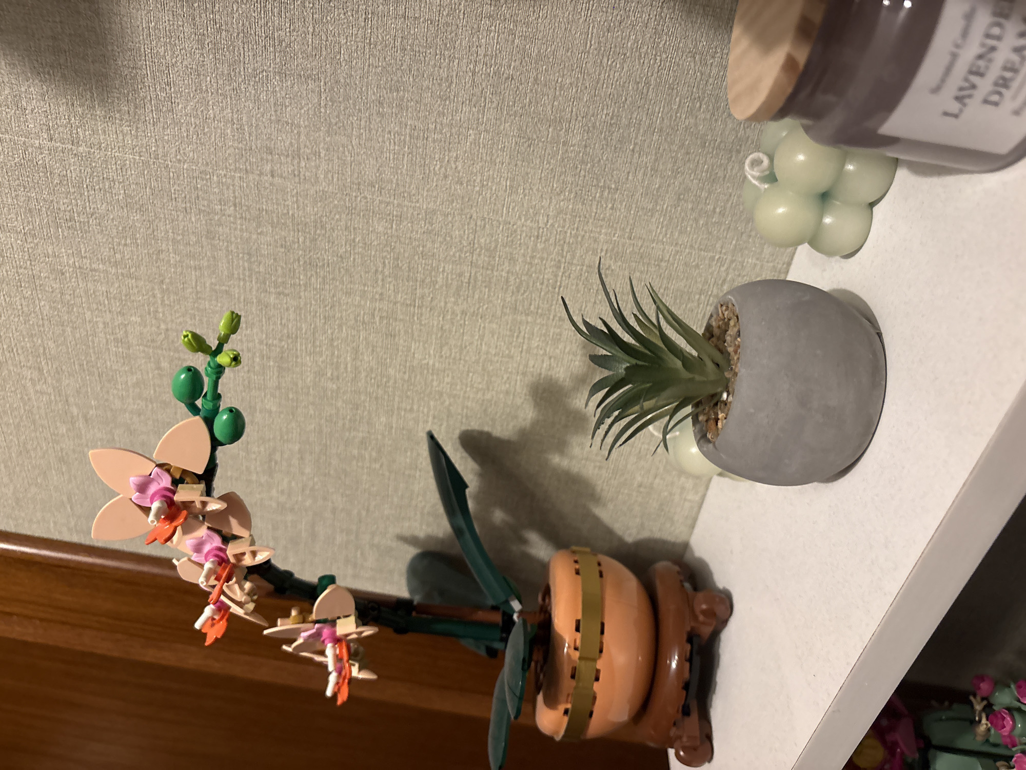 The image shows a shelf with several objects on it. A Lego orchid in a brown Lego pot is on the left. A small succulent in a gray pot and a bubble candle are next to it. A scented candle in a gray jar labeled "LAVENDER DREAM" is on the right. The objects are placed on a white shelf in front of a textured beige wall. There is no evidence to suggest the location is Sofia, Bulgaria.