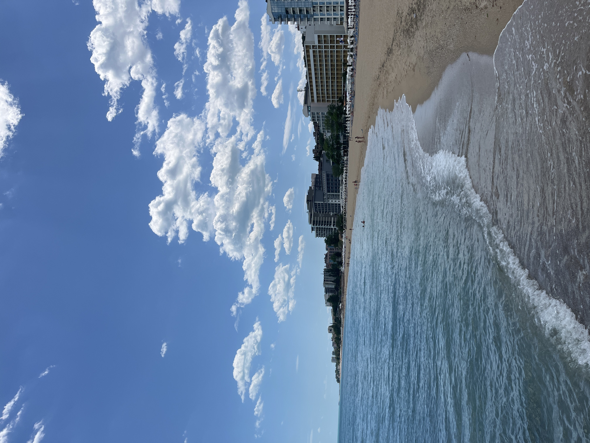 The image shows a beach scene in Varna, Bulgaria, with a clear blue sky filled with puffy white clouds. The beach stretches along the coastline, with light brown sand meeting the blue-green water. Gentle waves are visible near the shore. Tall buildings line the beachfront, suggesting an urban setting. People are present on the beach.