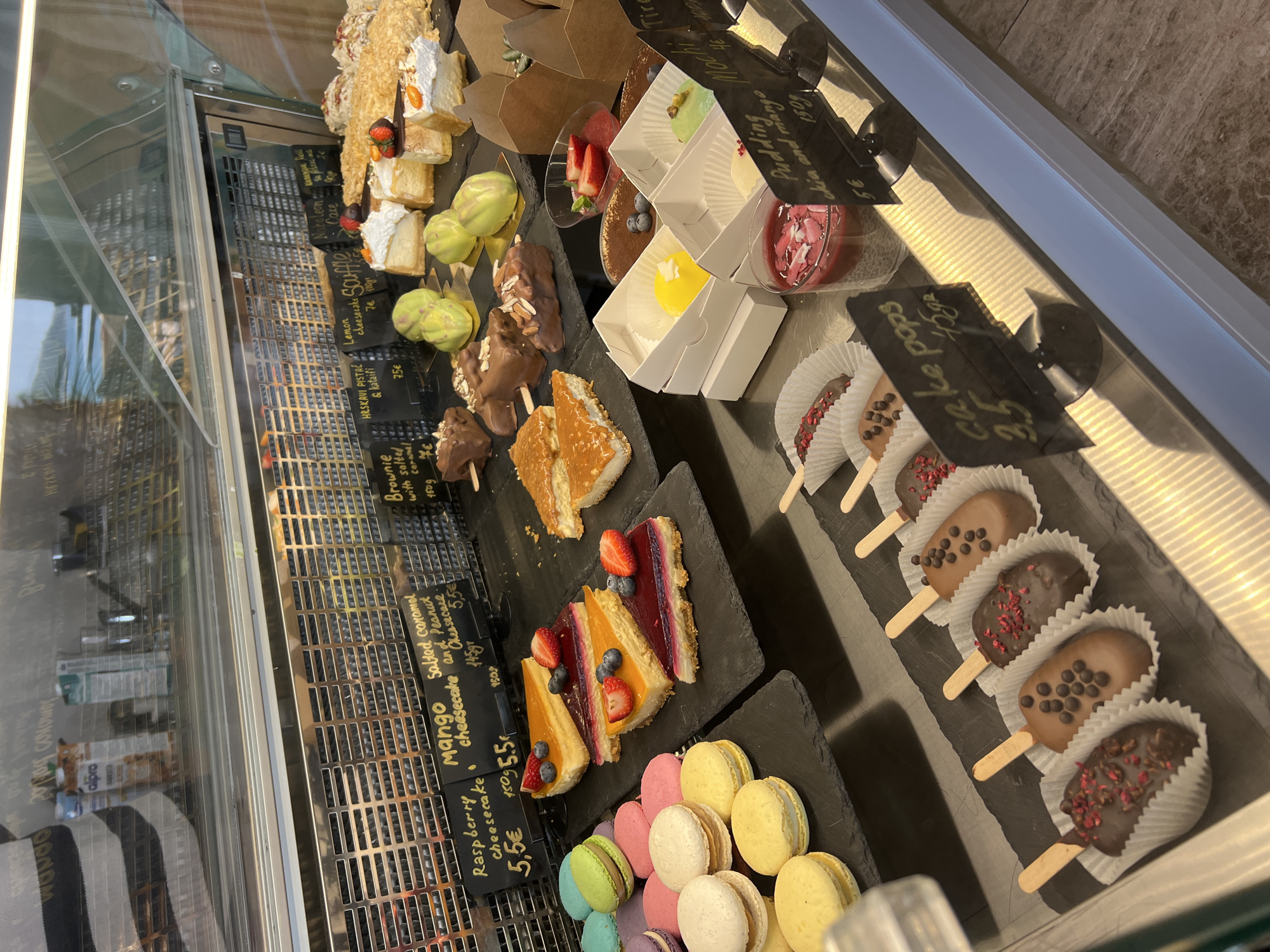 The image shows a display case in Rafailovići, Montenegro, filled with various desserts. The case is organized with different sweets arranged in rows. On the left are layered cakes, lemon cheesecake, and pistachio desserts. Moving to the right, there are brownies, cheesecakes (raspberry, mango, and salted caramel), macarons, puddings and mochi, and chocolate-covered cake pops on sticks. Price tags with descriptions are placed next to each item. The background shows a reflection of the outdoor scenery in the glass case.
