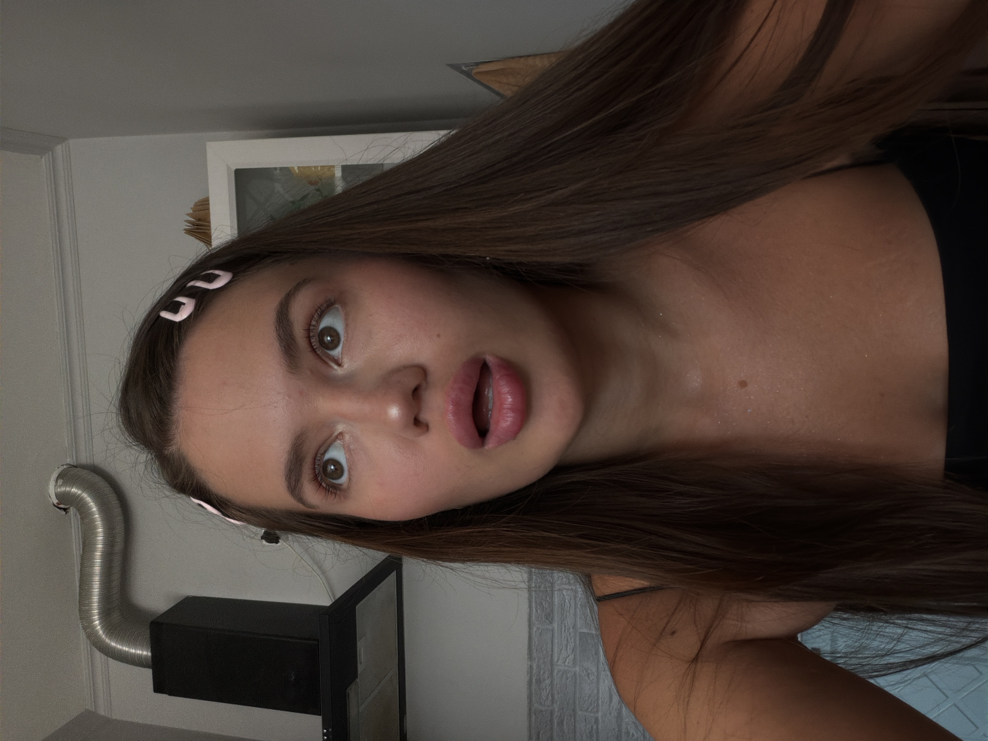 The image shows a close-up of a young woman with long, dark brown hair and light brown eyes. She is looking upwards and to her left with her mouth slightly open, suggesting surprise or curiosity. She is wearing a black spaghetti strap top, and two light pink barrettes are visible in her hair. The background appears to be an indoor domestic setting, possibly a kitchen, with a black exhaust hood and a silver ventilation pipe visible on the left. A white cabinet is partially visible on the right. The lighting suggests it is daytime, and the woman's skin appears slightly shiny, possibly from sweat. No specific details about the location in Sirius, Russia, can be determined from the image.