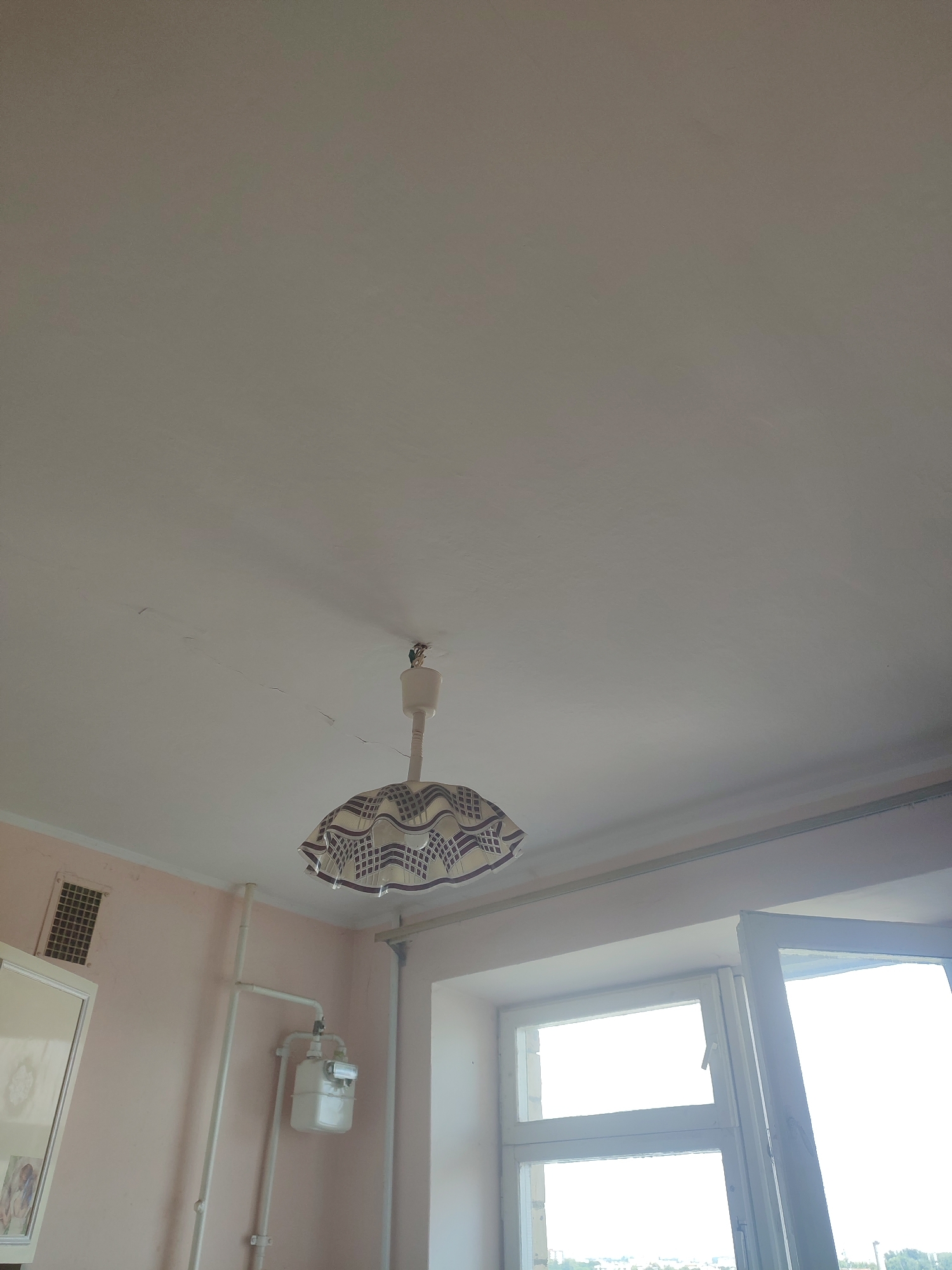 The image shows a ceiling in a room with a hanging light fixture. The light fixture has a decorative shade with a pattern of dark squares and circles on a light background. Cracks are visible on the ceiling. To the left of the light fixture, a white pipe with a gas meter attached is visible against a light pink wall. A ventilation grate is also present on the wall. To the right, a window with multiple panes is open, revealing a bright sky and a distant cityscape. The overall impression is of an interior scene in an apartment or house. The location is not specified in the image itself, nor can it be inferred from the visual information provided. Therefore, it is not possible to determine if the scene is in Chernivtsi, Ukraine.