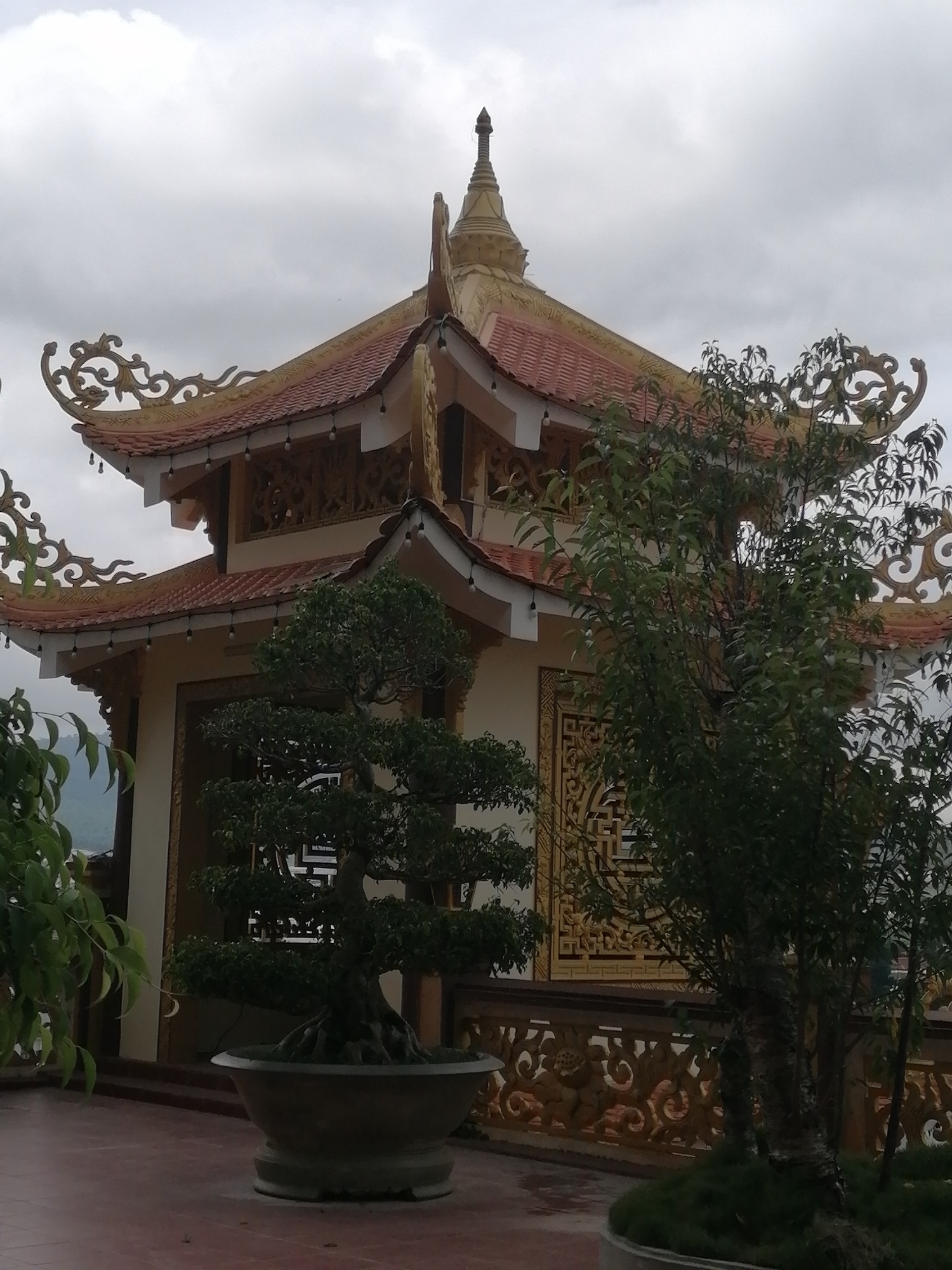 The image shows a multi-tiered, ornate building with a pagoda-style roof, typical of Vietnamese temples, located in the city of Lạc Dương, Vietnam. The building features red tiled roofs with decorative golden trim and intricate carvings. In the foreground, a large bonsai tree in a wide, shallow pot sits on a reddish-brown paved area. Behind the bonsai, a decorative golden fence with relief carvings is visible. To the right of the building, another tree with dense green foliage partially obscures the structure. The sky is overcast with heavy clouds. The overall scene depicts a serene temple courtyard with traditional Vietnamese architectural elements and carefully manicured greenery.