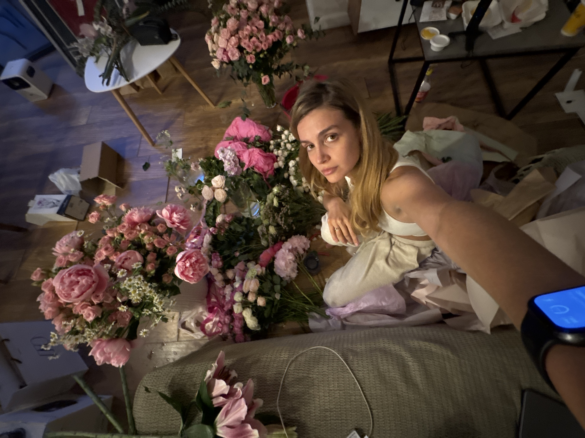 The image shows a woman sitting among a large arrangement of pink and white flowers on what appears to be a sofa or cushion. The flowers vary in type, including roses, peonies, and lilies. She is wearing a white top and light-colored pants. She is facing the camera, holding up her arm, on which she wears a smartwatch with a bright blue screen. Around her are discarded papers and packaging, suggesting that she may have been unpacking or arranging the flowers. In the background, there is a small white table, a black metal shelf with items on it, and scattered boxes and shopping bags, indicating an indoor setting. The location is unspecified.