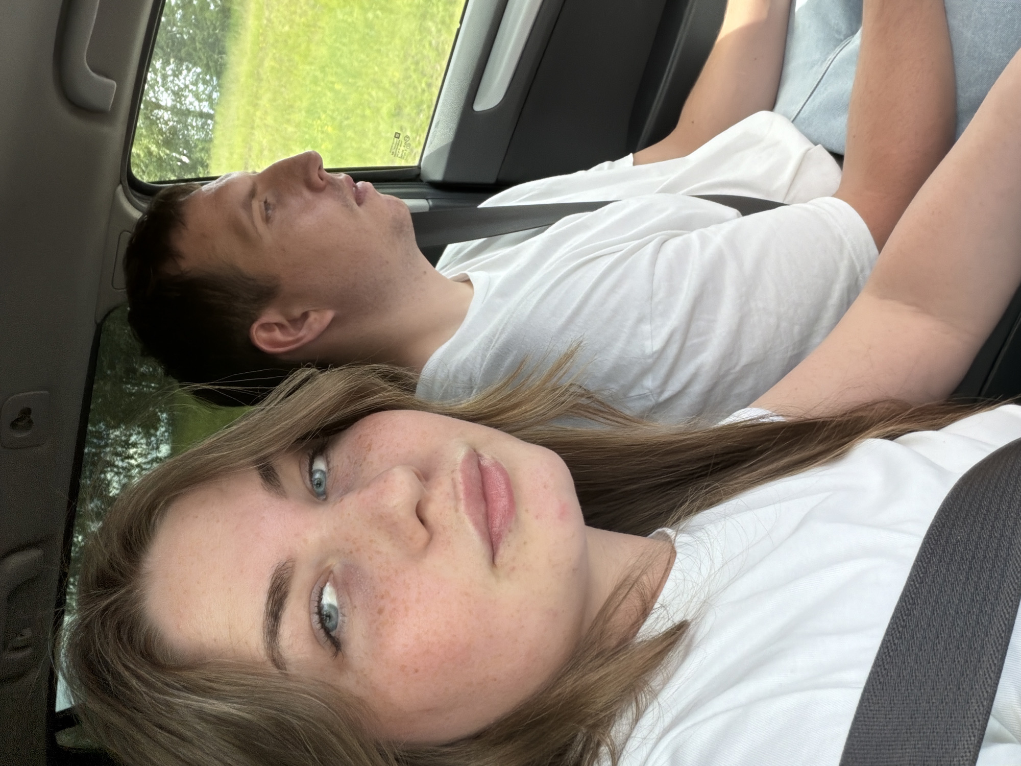 A young woman with long brown hair and blue eyes, and a man with short brown hair and brown eyes are sitting in the front seats of a car. The woman is in the foreground, looking at the camera, with her seatbelt across her chest. The man is in the background, looking out the window. Both are wearing white t-shirts. The car is moving, as indicated by the blurred greenery outside the window, suggesting a rural or suburban setting. The image does not provide any specific details about the city of Alnashi, Russia.
